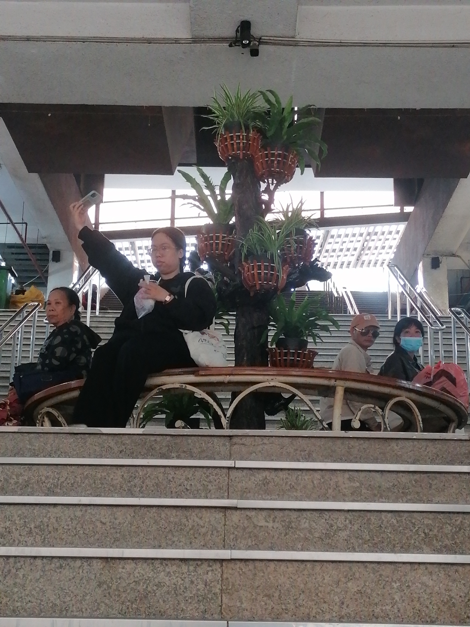 This image captures an indoor scene within a public space in Da Lat, Vietnam, likely a market, terminal, or large building. Four individuals are seated on a curved, ornate bench that encircles a decorative plant arrangement.
In the foreground center, a young Asian woman, dressed in a black long-sleeved top and pants, is actively taking a selfie. Her right arm is extended upwards, holding a smartphone, while her left hand holds a small plastic bag. A white tote bag with the word "ZARA" visible on its side rests beside her on the bench. To her left, an older Asian woman in a dark, patterned top sits composedly, looking towards the left side of the frame.
To the right of the central plant feature, an Asian man wearing a light-colored baseball cap and dark sunglasses sits, looking towards the viewer. Beside him, an Asian woman in a dark jacket wears a light blue surgical mask, also looking towards the viewer. A red bag is placed near her.
The central element of the seating area is a short tree trunk from which several potted plants, including spider plants and ferns, are arranged in woven basket planters. The bench itself features white, scrolled metalwork and a wooden top.
The background reveals multiple flights of light-colored stairs with metal railings, ascending to higher levels. A bright, patterned ceiling or window structure allows natural light into the space, indicating it is daytime. The immediate foreground shows several granite steps with metal anti-slip strips, leading up to the bench area. The scene depicts people resting and one engaging in a common modern activity, a selfie, within a clean, functional public environment.