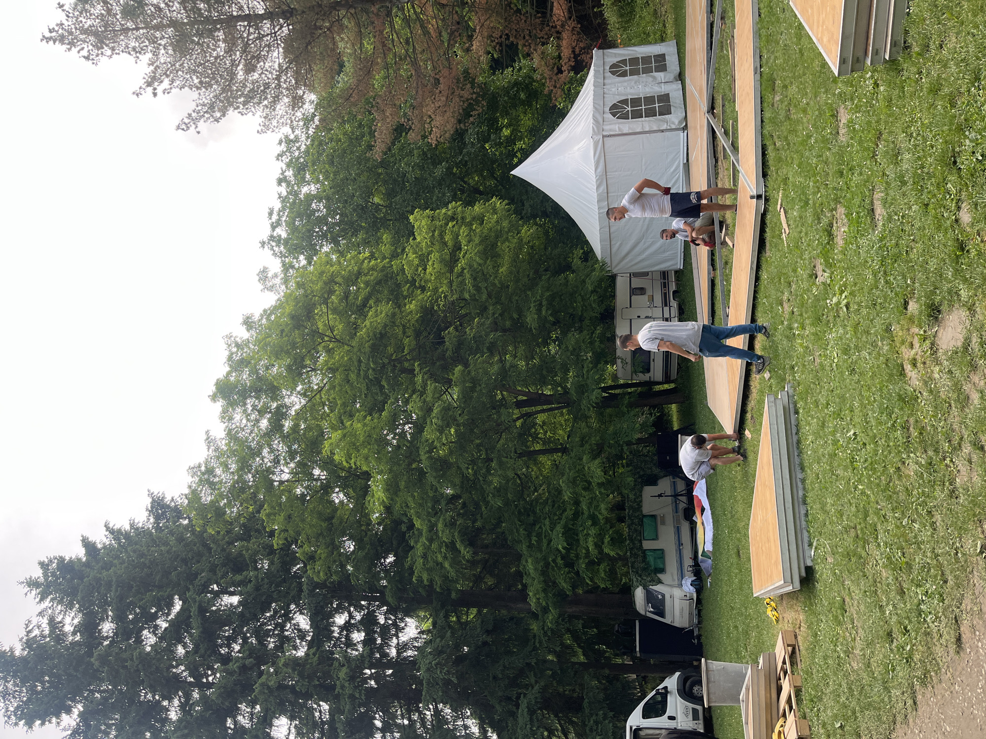 The image captures an outdoor scene in Sremski Karlovci Municipality, Serbia, depicting the setup for an event during daytime. Several men are actively engaged in assembling a temporary stage or flooring structure on a grassy field.
In the mid-ground, a large white event tent, featuring arched window panels, is set up. To the left of the tent and extending into the background, multiple camper vans or RVs are parked, alongside a white utility van in the foreground. The area where the men are working is covered with numerous wooden or plywood panels, laid out on metal framing to form a pathway or stage. More uninstalled panels are stacked nearby on the grass.
A man in a white t-shirt and dark shorts stands near the white tent, observing the progress. Closer to the foreground, another man in a grey t-shirt and blue jeans walks on a section of the constructed flooring. Further along the structure, a man in a white t-shirt kneels, appearing to work on the assembly.
The background is dominated by a dense treeline, suggesting a wooded or park-like environment. The sky above, visible in the upper left, appears overcast. The overall impression is one of active preparation for an outdoor gathering or festival. No discernible text is visible on any objects or signs in the image.
