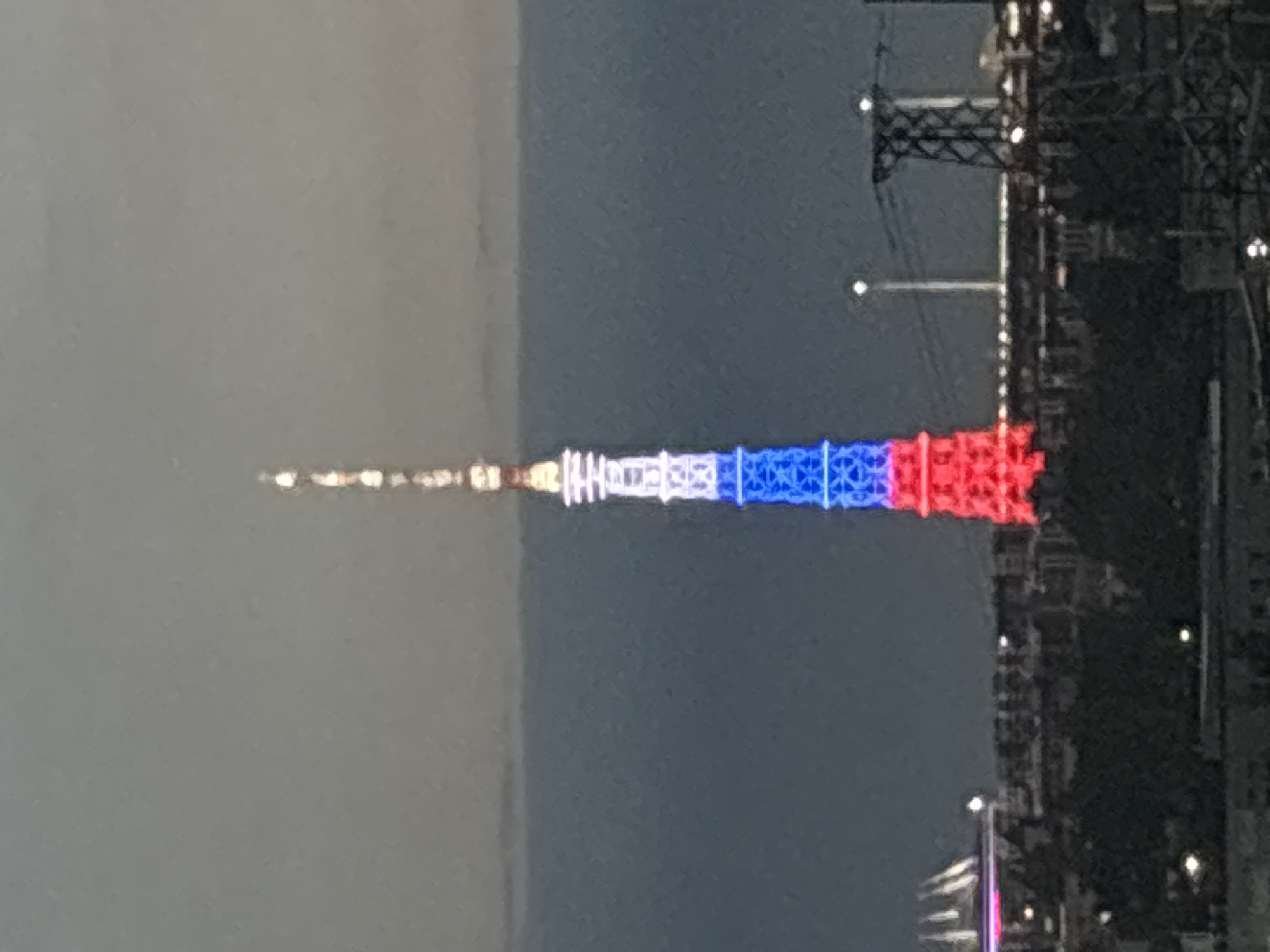 A telephoto, outdoor, daytime, landscape photograph captures a tall tower illuminated with red, white, and blue lights, resembling the colors of the Russian flag. The tower stands prominently in the center of the frame, with its peak extending into the overcast sky. Below the tower, a cityscape unfolds, characterized by dark, silhouetted trees and buildings. In the background, the outlines of more structures and infrastructure, possibly including power lines and a stadium-like building with pink lighting on the left, are visible against the dusky atmosphere. The image has a slightly blurred quality, suggesting it was taken from a distance or in low light conditions.