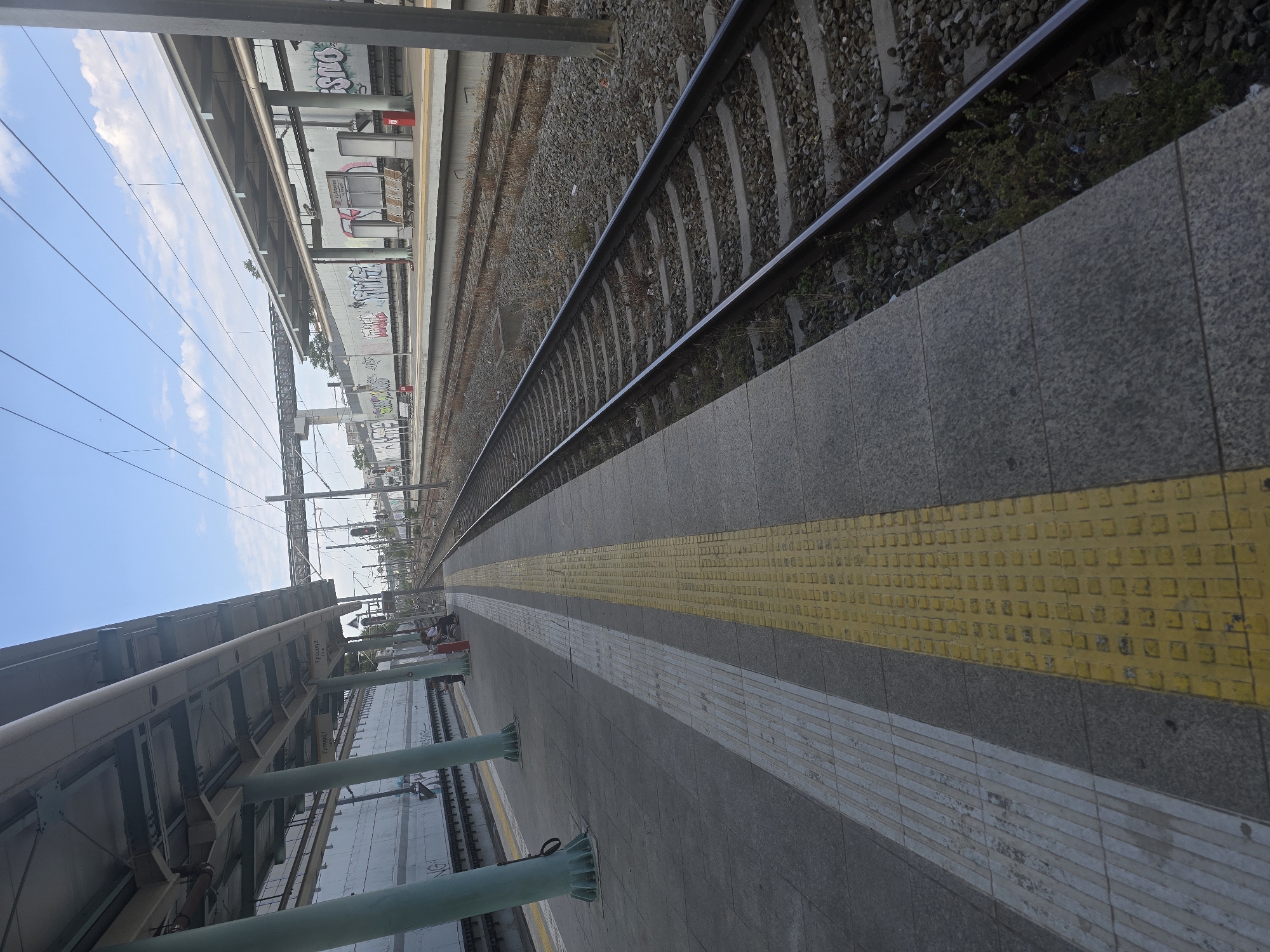 The image shows an outdoor view of a train station platform in Agii Anargiri, Greece. The platform is paved with grey tiles and features a yellow tactile paving strip along the edge closest to the camera, followed by a white tactile strip. Several green pillars support a covered roof over the platform, which has advertisements and graffiti visible on the wall behind it. To the right of the platform, train tracks are laid on a ballast of grey stones, with some weeds growing between the sleepers. The tracks extend into the distance under a bright blue sky with scattered white clouds. In the background, there are overhead power lines and structures associated with the railway. A few people are seated on benches on the platform in the mid-ground, waiting.