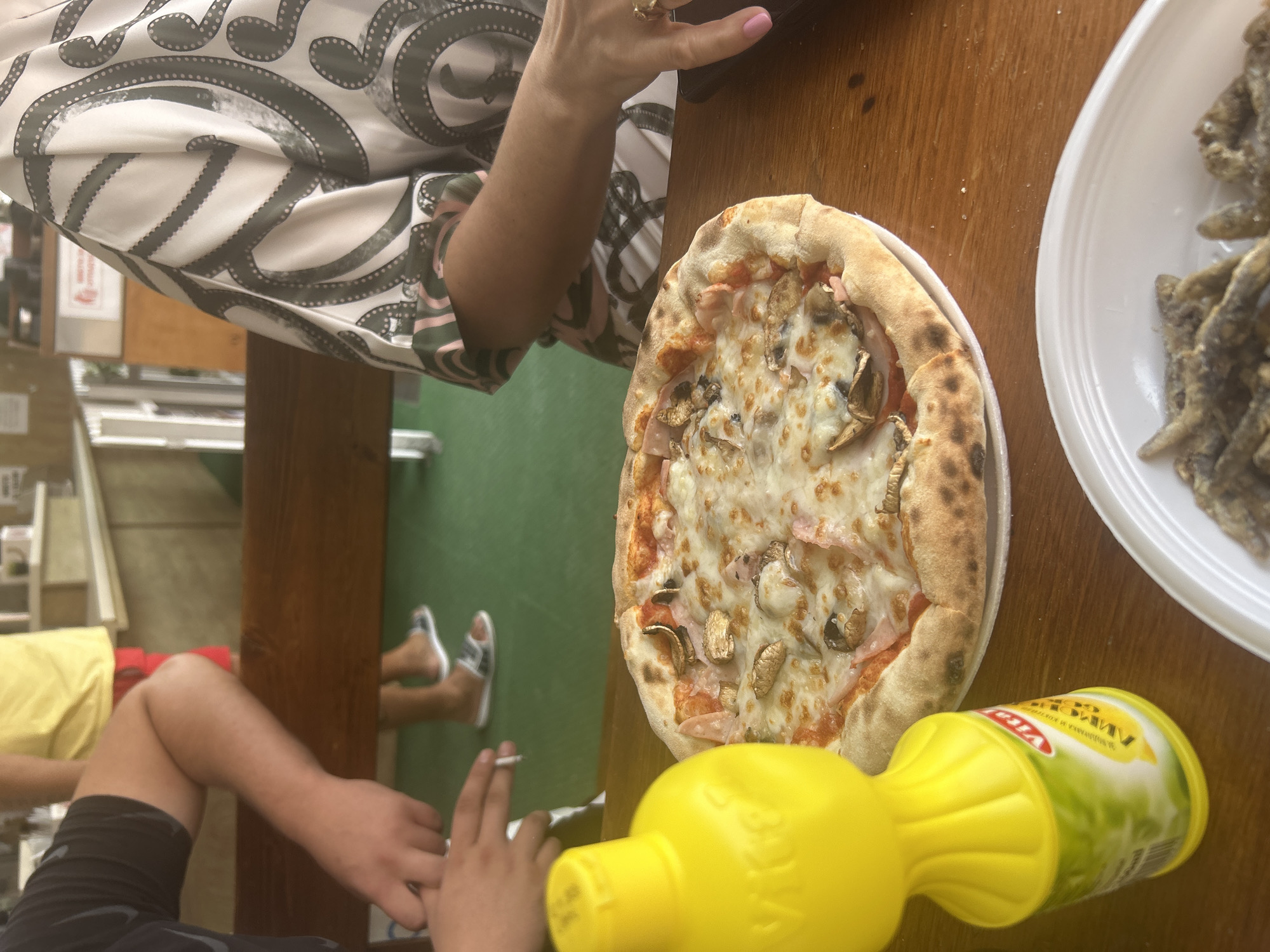 In this close-up, eye-level shot, a table is laden with food and beverages. In the foreground, a bright yellow squeeze bottle with a red and white label that reads "Vita" is positioned to the left. To the right of the bottle, a circular pizza rests on a white plate, topped with melted cheese, mushrooms, and ham. A white plate filled with small, fried fish is visible at the bottom right of the frame. The background reveals a green floor and a wooden bench, with the legs of two people wearing casual attire and flip-flops. A hand holding a lit cigarette is seen in the upper left quadrant, leaning on the bench. The upper portion of the image shows a person wearing a white shirt with a black and gray intricate pattern. The scene appears to be set in an outdoor dining area.