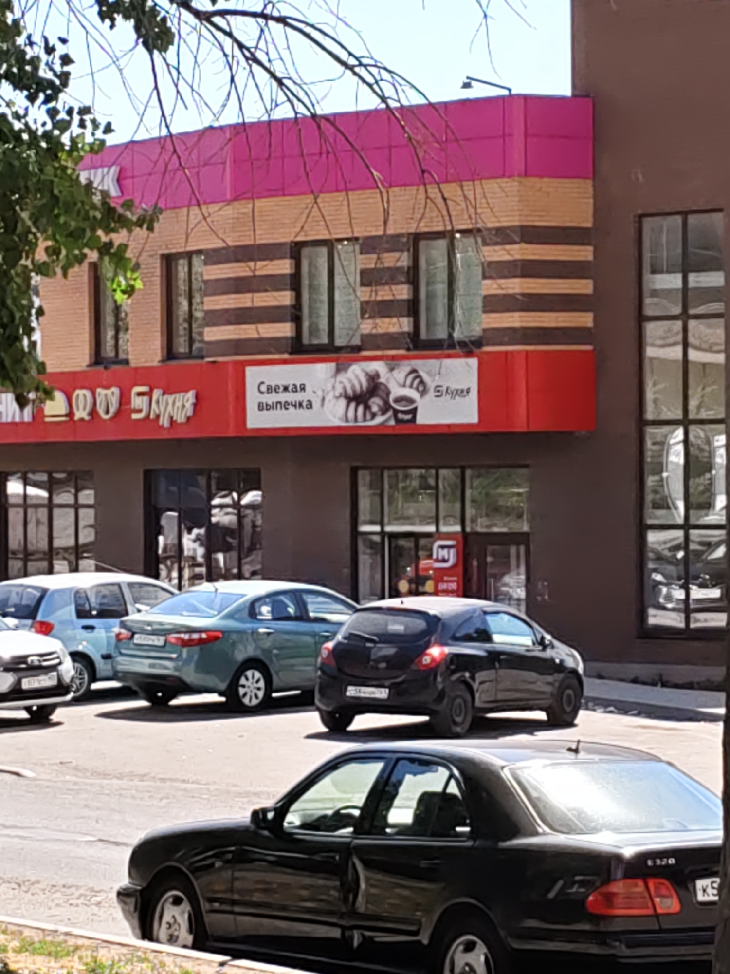 The image depicts a street scene in front of a commercial building in Rostov-on-Don, Russia. The building features a modern facade with pink and striped sections, and large windows. A red sign above the entrance displays Cyrillic text, with one part appearing to be "Кухня" (Kitchen) and another smaller sign with an image of pastries and coffee stating "Свежая выпечка" (Freshly baked goods). Several cars are parked on the asphalt road in front of the building. These include a light blue hatchback, a green sedan, a black hatchback, and a black sedan in the foreground. Trees with green foliage are visible on the left side of the image, and the sky is clear. The overall impression is a typical urban commercial street view.