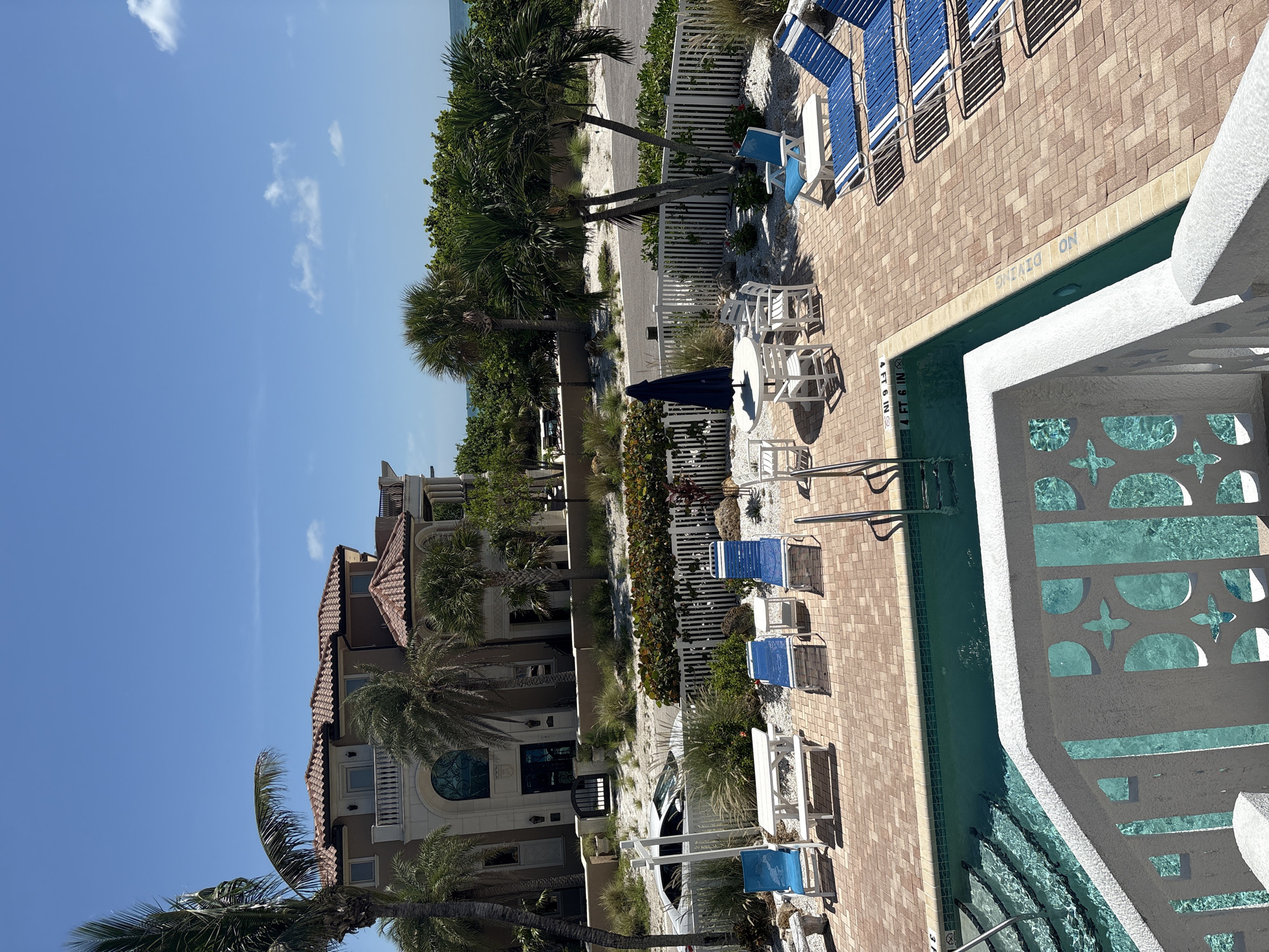 This image depicts a view of a swimming pool area and a luxury beachfront property in Holmes Beach, Florida, United States of America. The scene is set on a sunny day with a clear blue sky and a few scattered clouds.
In the foreground, a swimming pool with clear turquoise water is visible, featuring a set of stairs leading into it. A white railing with decorative cutouts partially obscures the pool. The pool deck is paved with reddish-brown brick pavers.
Several blue lounge chairs with white frames are arranged on the deck. Some of these chairs are empty, while others appear to be occupied by unseen individuals. A white picnic table with benches sits near the pool's edge, and a white outdoor table with chairs is also present, accompanied by a closed blue umbrella.
Beyond the pool area, a white picket fence separates the property from a sandy area with vegetation and palm trees. A large, multi-story, Mediterranean-style house with a terracotta tile roof dominates the background, suggesting a high-end residential or resort property. A white car is parked in the driveway in front of the house. The overall environment suggests a tropical, resort-like setting, typical of coastal Florida.