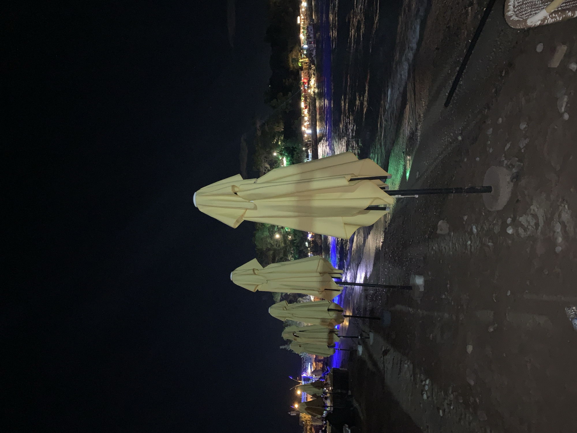 The image depicts a beach at night in Kemer, Turkey. The foreground is a dark, pebbly beach with several closed and covered beach umbrellas lined up along the shore. The umbrellas are a light yellow or cream color. In the background, the sea is visible, with gentle waves lapping the shore. Across the water, a strip of lights indicates a town or resort area with buildings and possibly restaurants. The sky is dark and clear, typical of a night scene. There are no people clearly visible in the immediate foreground. The overall atmosphere is calm and serene, suggesting the end of a day at the beach.