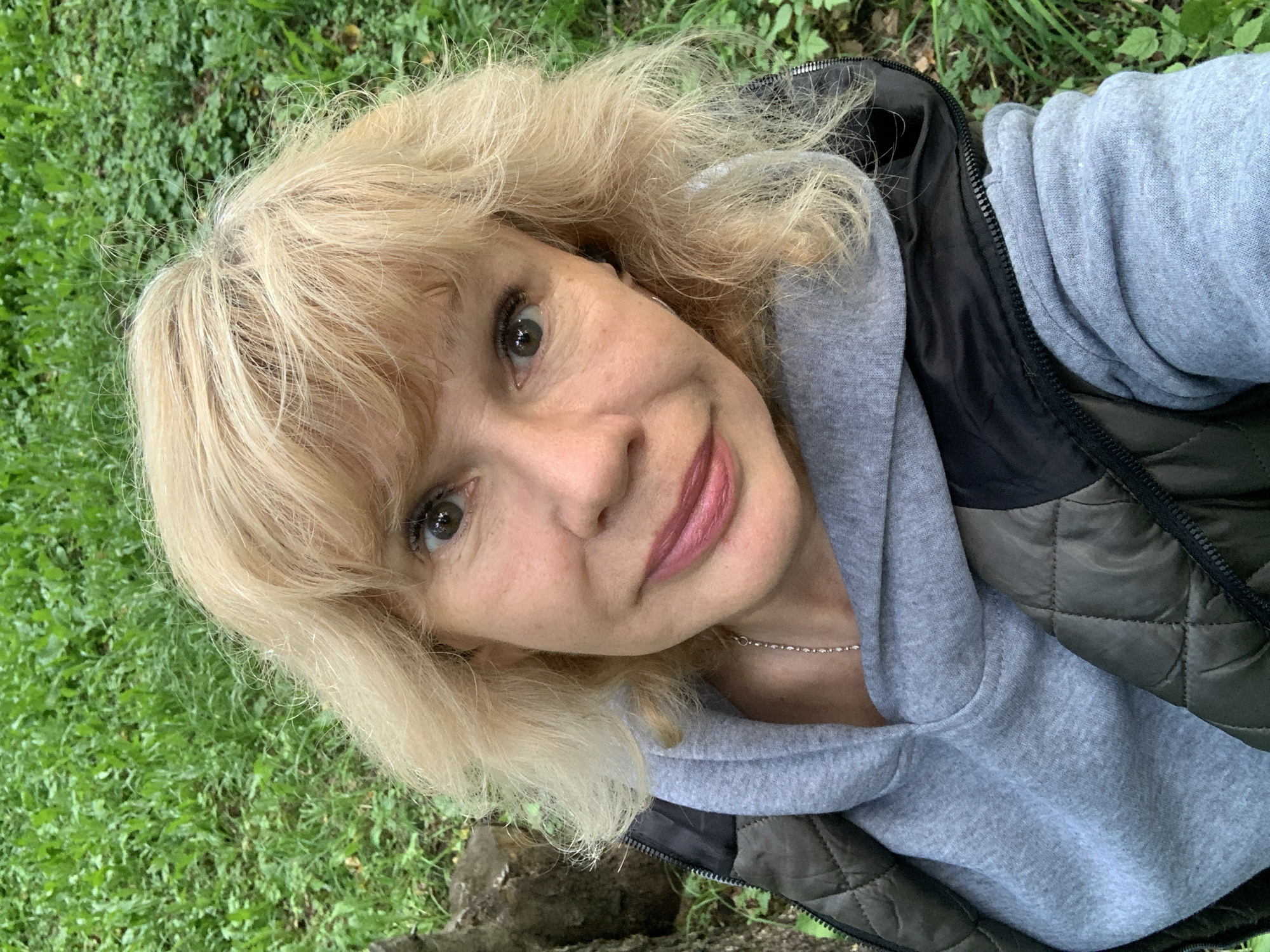 A close-up, high-angle selfie shows a fair-skinned woman with blonde, wavy hair. She is wearing a grey hooded sweatshirt and a dark green quilted vest. She is smiling gently, looking directly at the camera. The background consists of green grass and foliage, suggesting an outdoor setting. The image was taken in Prokopyevsk, Russia.