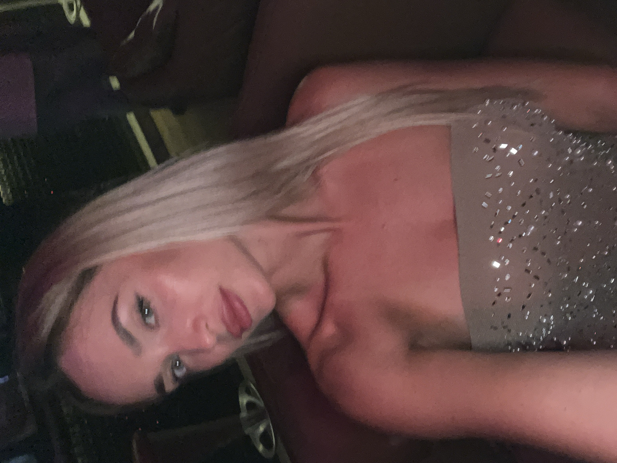 The image shows a close-up selfie of a young Caucasian woman with blonde hair. She is wearing a strapless, embellished dress. The lighting suggests an indoor setting, possibly a restaurant or club, with warm, reddish-pink tones dominating the scene. The background is dark and out of focus, with some hints of dark furniture. There is no discernible indication that this image was taken in Sharm El Sheikh, Egypt.