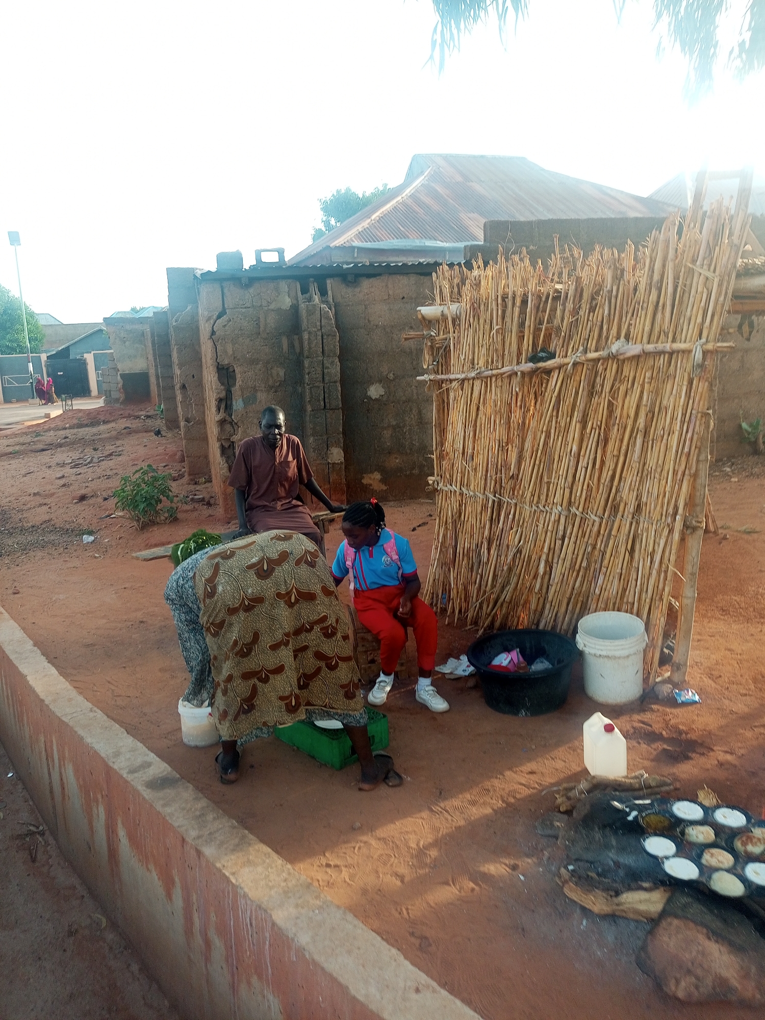 The image shows an outdoor scene in a residential area. Three people are visible: an elderly man in brown clothes sitting on a bench next to a young girl in a school uniform, and an elderly woman in a patterned dress bending over. Near them are a bamboo screen, a black bucket, a white bucket, and a container. In the foreground, food is being cooked on a small stove. The ground is reddish-brown. Buildings with corrugated roofs and dilapidated brick walls are in the background. The location is the city of Unknown, Unknown.