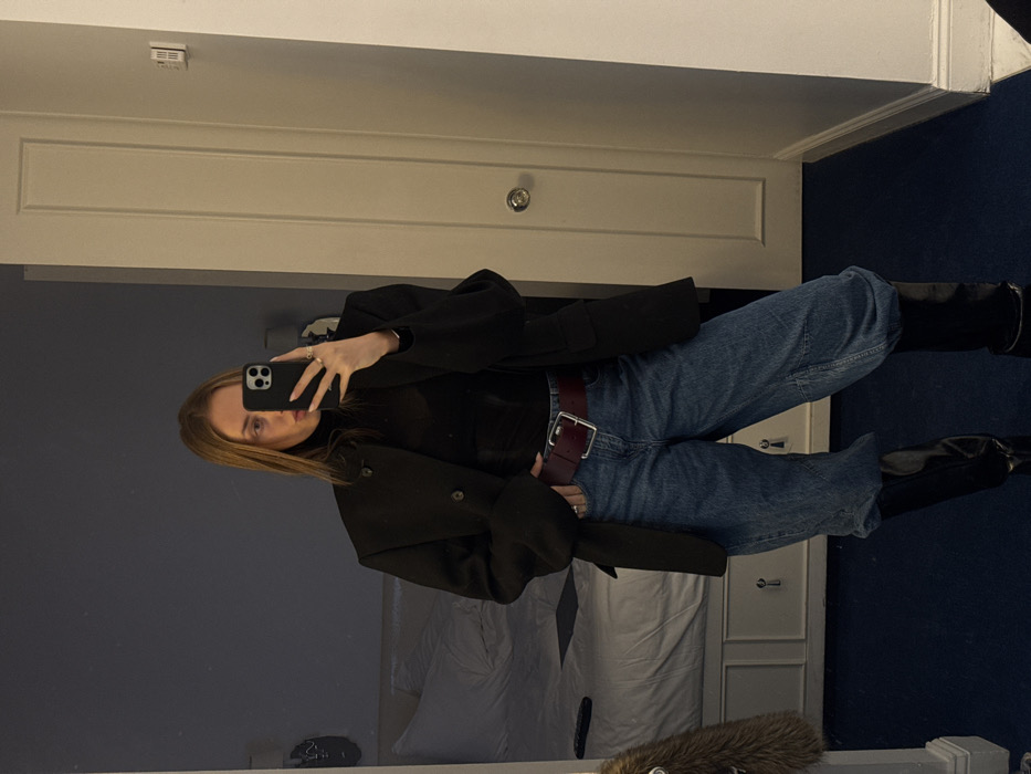 A person stands in front of a mirror, holding a phone to take a picture. They wear a black turtleneck, blue jeans secured with a brown belt, a black coat, and black boots. A bed with white linens is visible in the mirror’s reflection. The walls of the room are light blue and white. A dark blue carpet is present on the floor.