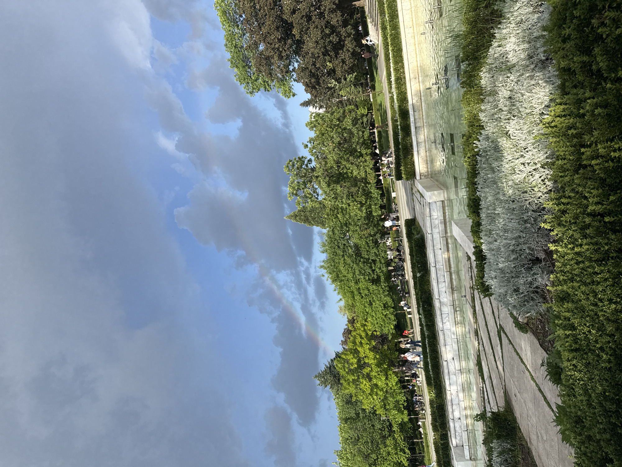 The image depicts a view of the city of Sofia, Bulgaria. The view is toward the sky, with a partial rainbow visible against a backdrop of clouds. Below the sky, there are trees and a stepped water feature or fountain lined with green hedges and silver shrubs. On the left side of the fountain, there are people walking and cycling alongside the green space.