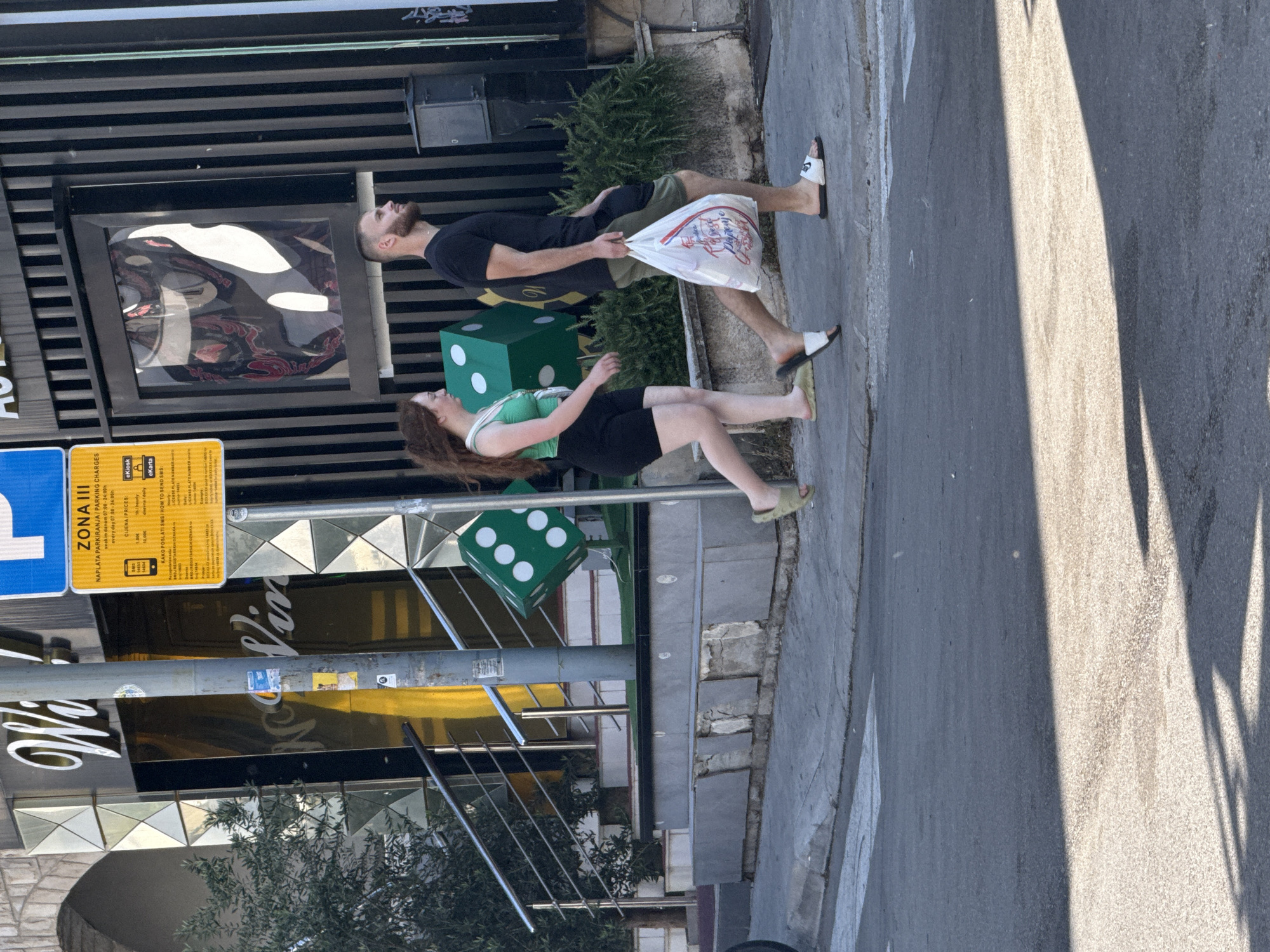 A young man and woman are walking on a sidewalk in Budva, Montenegro. The man is carrying a white plastic bag in his right hand. The woman is walking to his left and slightly behind him. They are walking past a building with large dice decorations in front of it. The building has a gold facade and a black striped exterior. There is a yellow sign with black lettering to the left of the dice. The sidewalk is cracked in places. The street in front of the sidewalk is paved with asphalt and has a pedestrian crossing marked with white lines. Sunlight casts shadows on the street.
