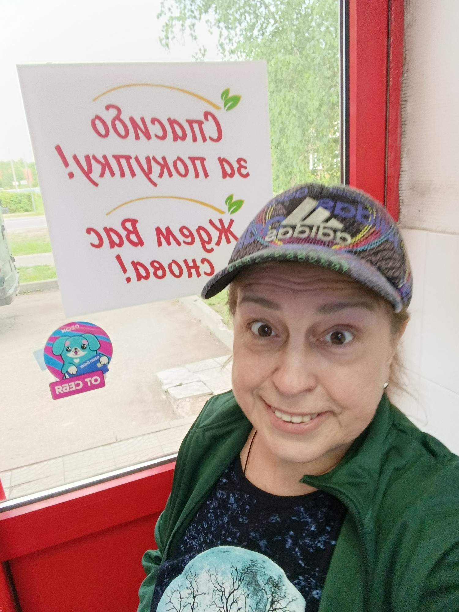 A woman wearing an Adidas baseball cap and a green zip-up jacket over a dark patterned t-shirt is taking a selfie. She has a surprised expression with wide eyes and a slight smile. The background shows a white sign with red Russian text, possibly indicating thanks or a message from a business. There is also a circular sticker with a cartoon dog on the sign. The scene appears to be outdoors, with trees and a street visible through a window to the left. The right side of the image shows a red frame, possibly of a building entrance or window. The setting is the city of Vereya, Russia.