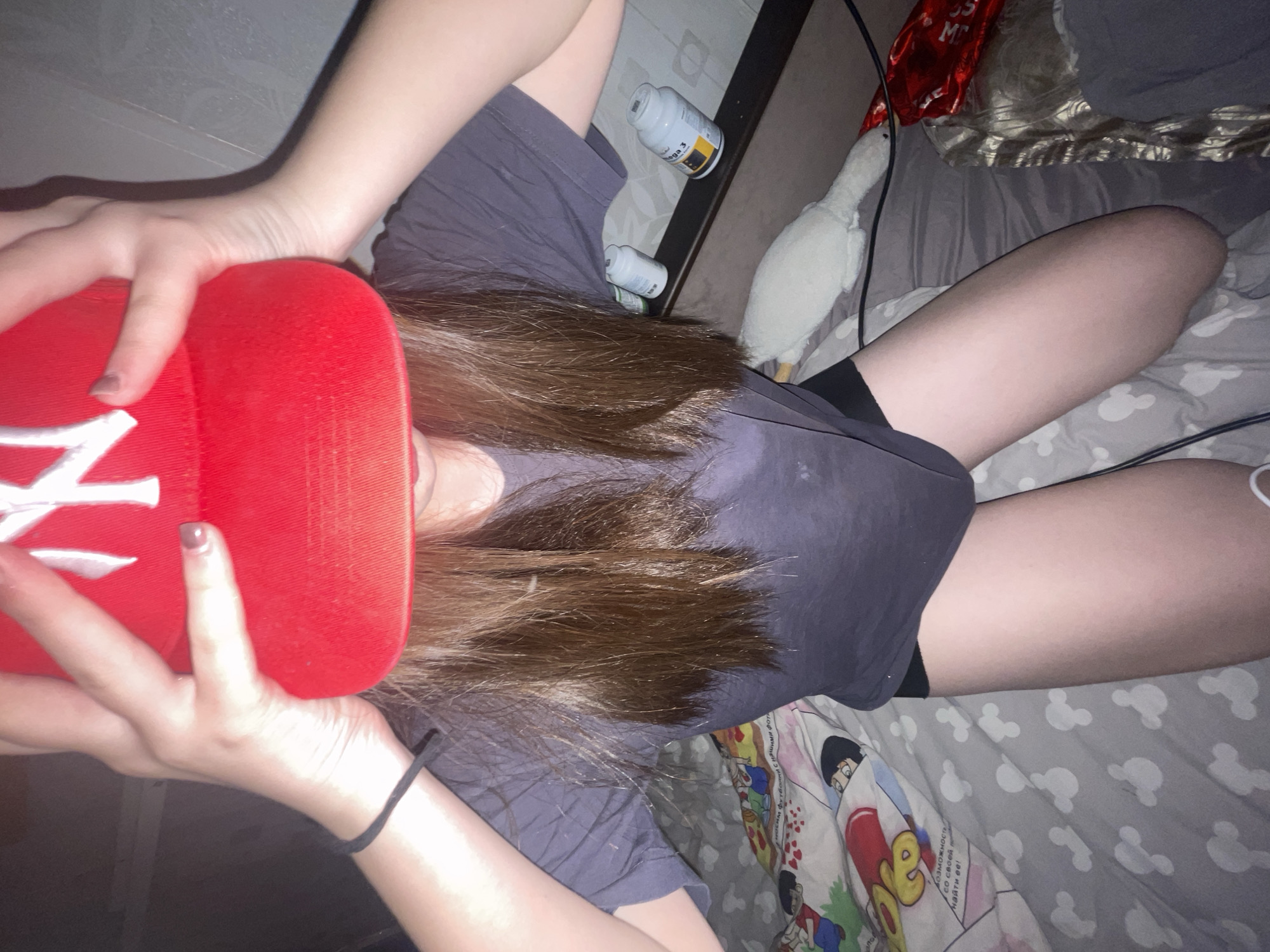 The image shows a person lying down, likely on a bed, in what appears to be an indoor setting in Murino, Russia. The person, who has long brown hair, is wearing a dark t-shirt and dark shorts or underwear. Their right hand is holding or resting on a red baseball cap with a white "NY" logo partially visible. A black elastic band or bracelet is on their left wrist, and their fingernails have dark polish.
Around the person, several objects are visible. On a surface next to them, there are two white bottles, one clearly labeled "Omega 3" and another with a visible "M". A white stuffed animal resembling a goose or duck is positioned near the person's legs. The bedding features various patterns, including grey fabric with white Mickey Mouse heads and a colorful sheet with cartoon characters and Cyrillic text. Visible Cyrillic phrases on the bedding include "М Футбол с нашими фот", "Козможность", "со своей п", and "найти ее!". The lighting is dim, suggesting it might be nighttime or a poorly lit room, contributing to a relaxed atmosphere. The person's posture indicates they are resting or relaxing.