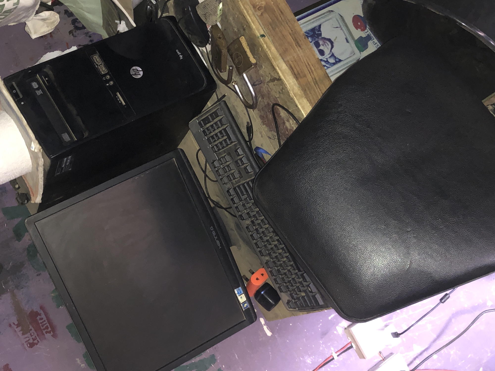The image shows a cluttered workspace with various objects. A black HP desktop computer tower is visible, along with a black computer monitor and keyboard. A black chair with a leather seat is positioned to the right. Other miscellaneous items are scattered around, including a wooden object, a lock, and several cables. The environment appears disorganized and potentially dirty. The city and location are not discernible from the image.