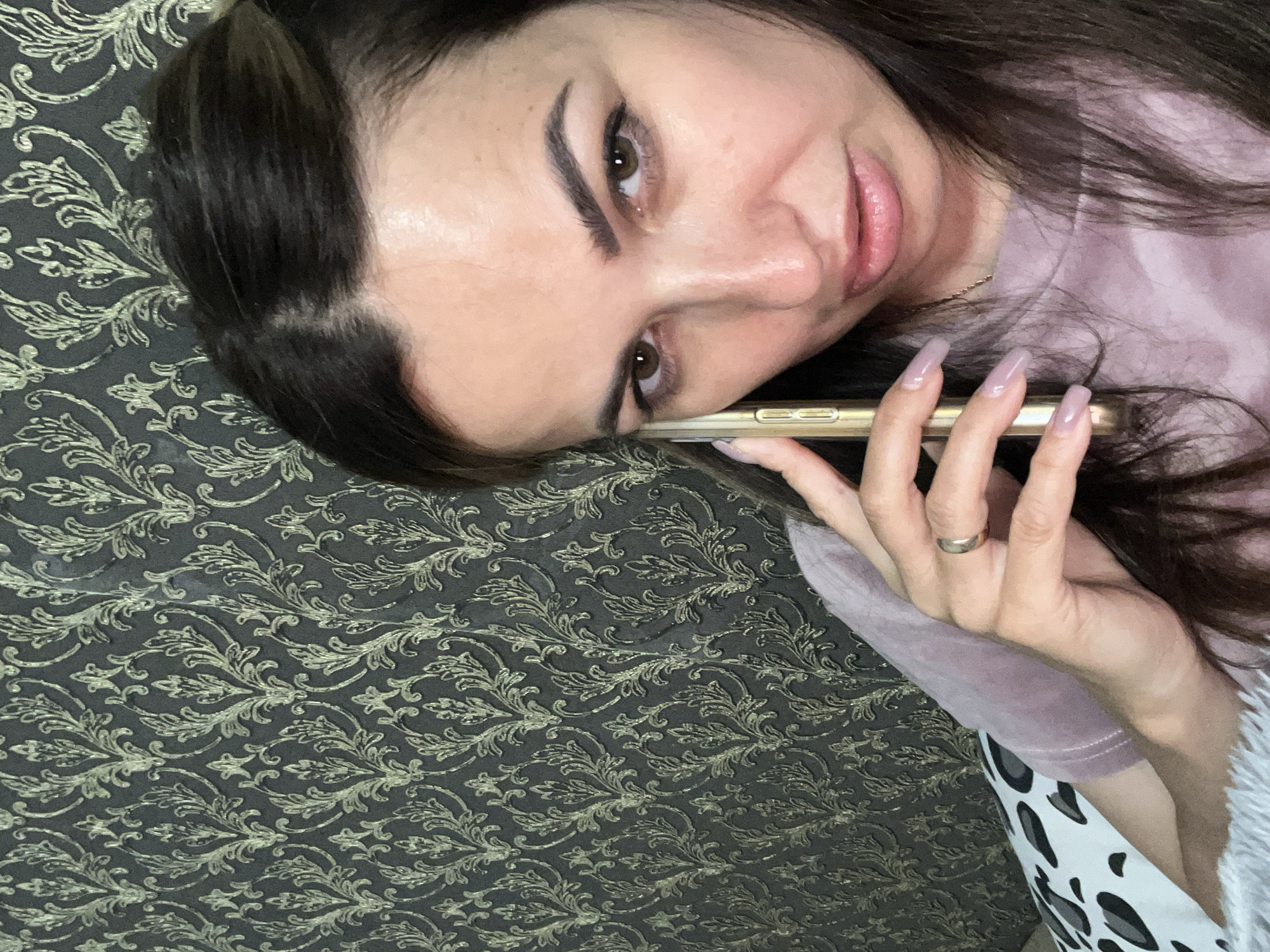 An overhead medium shot captures a woman with dark hair and brown eyes speaking on a gold smartphone. She is wearing a pink t-shirt, and her left hand, adorned with a silver ring, is holding the phone to her ear. The woman is positioned in the foreground, with her face filling most of the frame. Behind her, a dark gray wallpaper with a repeating ornate gold pattern covers the wall. The lighting in the image appears to be from above, casting subtle shadows on her face. The overall scene suggests an indoor setting.