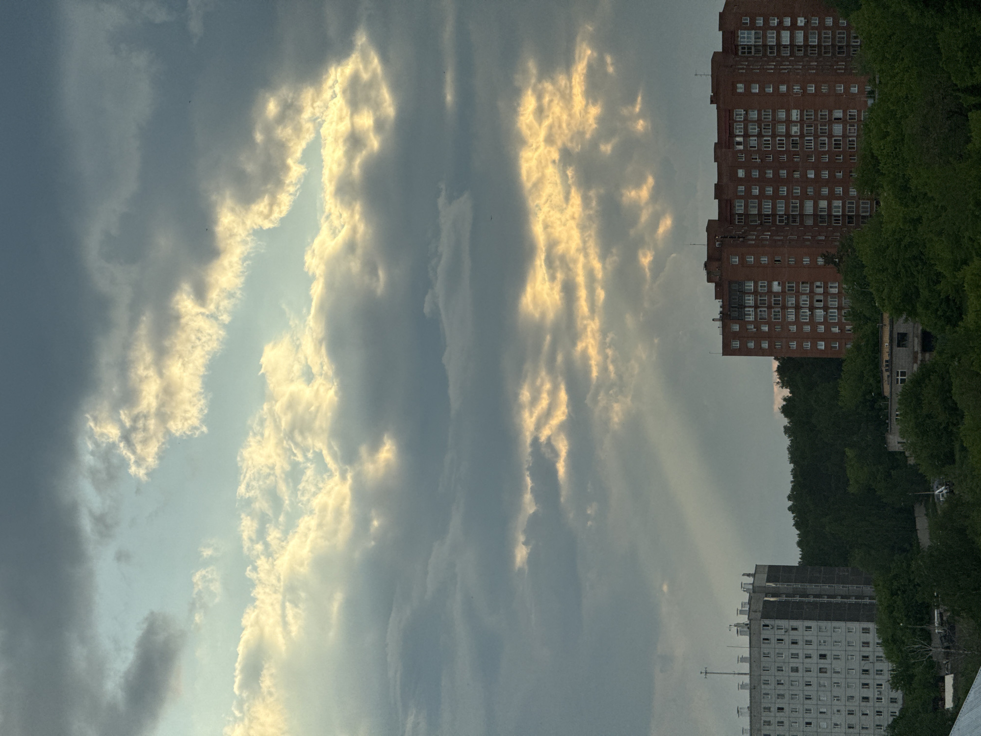 The image depicts a cloudy sky with shafts of sunlight breaking through, over a cityscape. In the foreground, a dense line of green trees forms the base of the scene. Behind the trees, two large apartment buildings are visible. The building on the left is a tall, gray, multi-story structure with many windows and visible antennas on its roof. The building on the right is a larger, red brick apartment complex, also with numerous windows, stretching horizontally across the frame. The sky above is filled with dramatic clouds, some dark and heavy, others illuminated by the sun, creating a dynamic interplay of light and shadow. The scene suggests a residential area within the city of Krasnoyarsk, Russia, likely captured during a transitional period of weather. There are no people or visible actions in the image.
