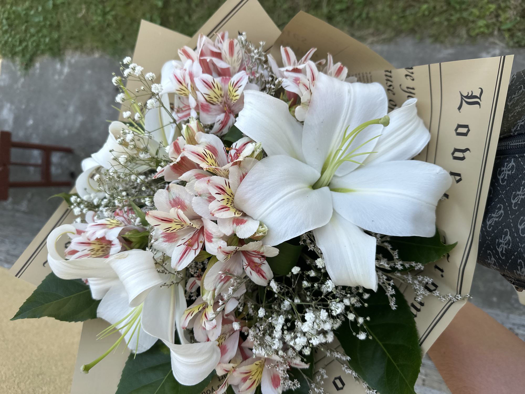 A close-up, eye-level, outdoor shot captures a bouquet of flowers, primarily white lilies and pink and white alstroemeria, artfully arranged and wrapped in brown paper with text. The bouquet is held by a person whose arm and shoulder are visible on the left. The background features a blurred outdoor setting with a concrete path or ground and green grass, suggesting a location in Marten, Bulgaria. The lighting is natural and diffused, highlighting the delicate textures and colors of the flowers. The overall composition is focused on the floral arrangement, with the background providing context.