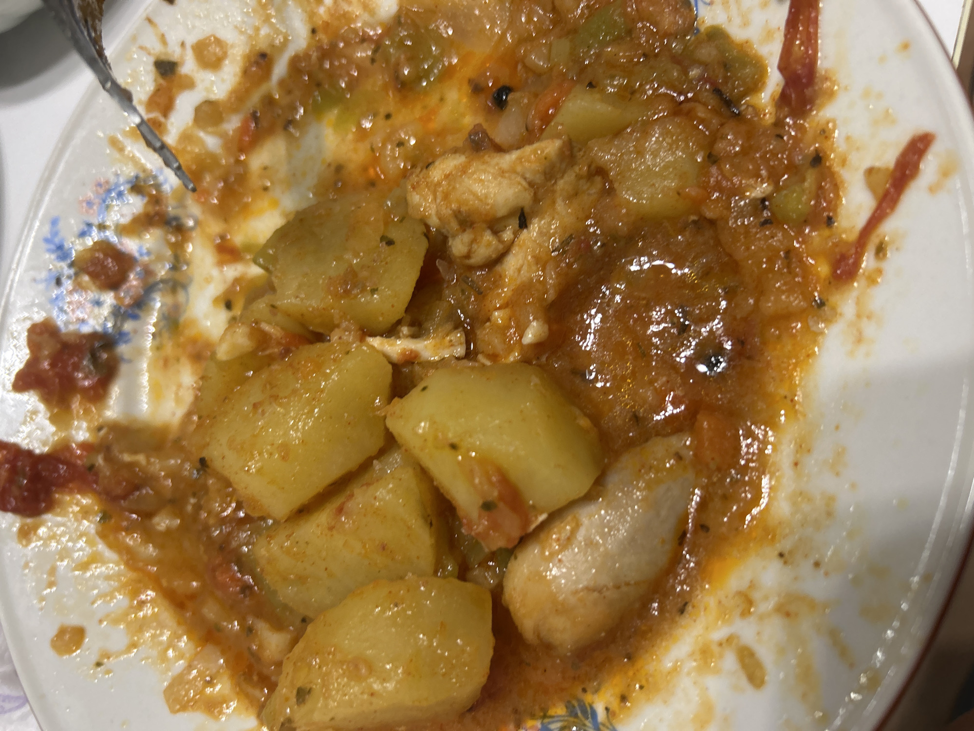 The image shows a close-up, overhead view of a white plate with a floral rim, containing a stew of potatoes and chicken in a reddish-orange sauce. The food is partially eaten, with a fork visible at the top right, suggesting a meal in progress. The setting appears to be indoors, possibly a restaurant or home, but no specific geographical details about Plovdiv, Bulgaria are present in the image.