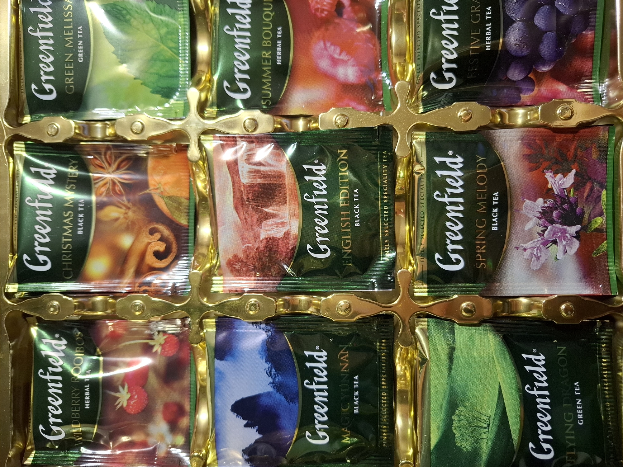 The image is a close-up, top-down view of a gift box containing individually packaged tea bags. The box is a metallic gold color with embossed decorative elements. The tea bags are arranged in a grid of three rows and three columns. Each tea bag features the "Greenfield" logo prominently, along with the specific tea flavor. The flavors visible are: Wildberry Rooibos, Christmas Mystery, Green Melissa, Magic Yunnan, English Edition, Summer Bouquet, Flying Dragon, Spring Melody, and Festive Grape. Each tea bag also displays an image related to its flavor, such as strawberries, cinnamon, green leaves, mountains, waterfalls, raspberries, a lone tree in a green field, flowers, and grapes. The overall presentation suggests a premium product. The scene is indoors, and there is no indication of the city of Tyumen, Russia, or any specific location.