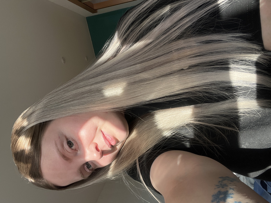 An indoor selfie depicts a light-skinned female individual in the city of Chaikovsky, Russia. She is shown from the chest up, positioned slightly to the left of the frame, looking directly at the camera with a slight smile. Her long, straight hair features darker roots blending into light ash blonde lengths, with strands falling over her shoulders. Strong, directional sunlight illuminates the right side of her face and hair, creating distinct patterns of light and shadow. She wears a dark, possibly black, short-sleeved top. A blue floral-patterned tattoo is visible on her left forearm. The background reveals a light-colored ceiling with two visible recessed lights in the upper right. Below the ceiling, a section of a teal-colored wall is visible, bordered by a wooden door frame or trim on the right.