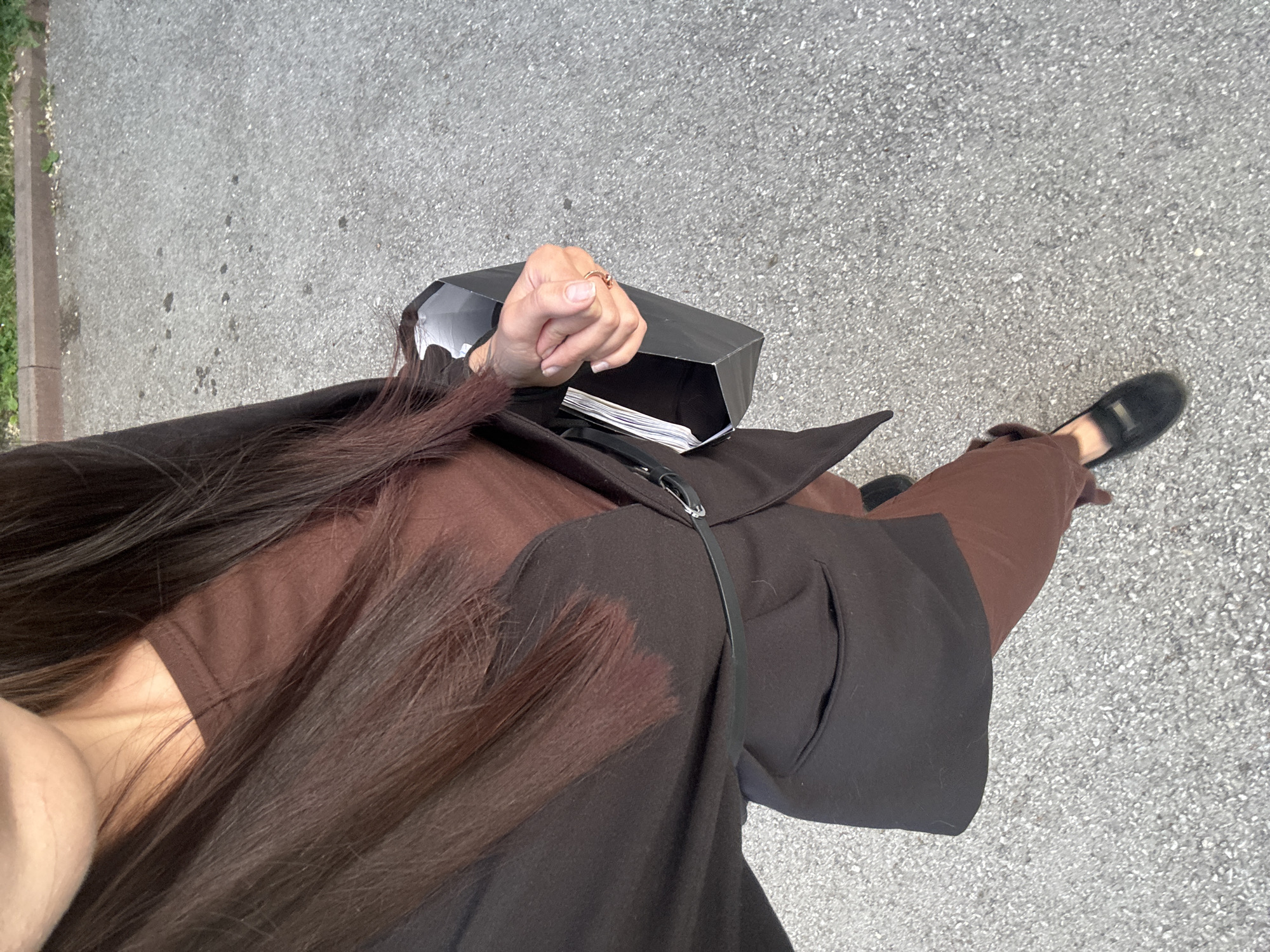 The image, taken from a high angle, shows a person from the chest down, standing on an asphalt surface with a grassy area and a curb in the background. The person is wearing a brown long-sleeved dress and a dark blazer cinched with a thin black belt. They are holding a black geometric bag in their right hand, which contains what appears to be papers. Their left hand is resting on their right arm. They are also wearing black loafers. The overall scene suggests an outdoor urban environment. The specific location within the city of Inskoy, Russia, cannot be determined from the visual information alone.