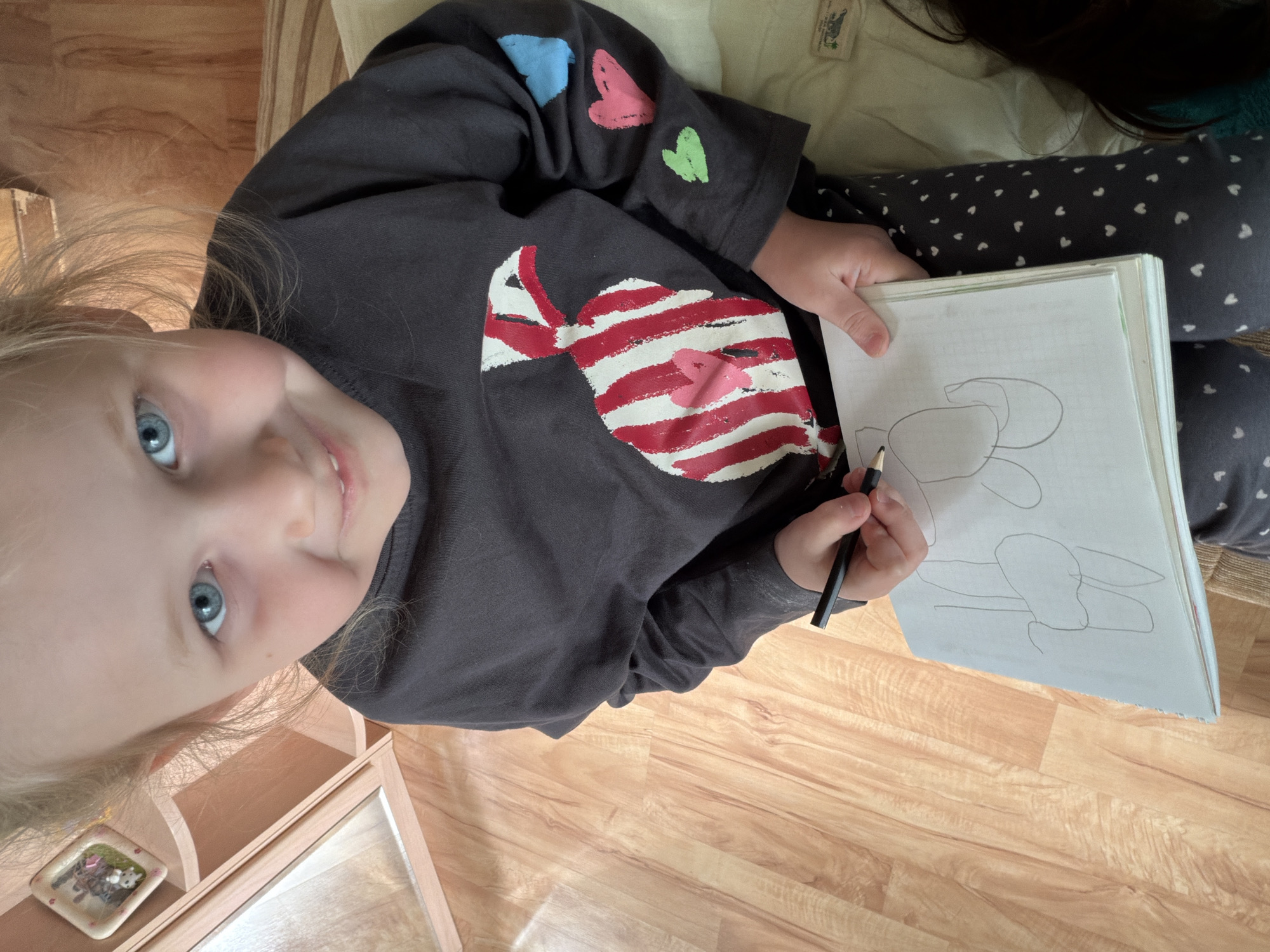 A young, fair-skinned child with blonde hair and blue eyes sits on a light-colored striped cushion or seat, holding a sketchbook and a black pencil. The child is smiling and looking towards the camera. The child is wearing a gray long-sleeved shirt with a red and white striped heart on the front and small colored hearts on the sleeve. The child is also wearing gray leggings with small white hearts. The sketchbook is open, showing a sketch of what appears to be an animal or character with rounded shapes. The background consists of a wooden floor, a wooden side table with a small decorative frame on it, and an off-white cushion or surface. The image was likely taken indoors. There is no information in the image that mentions Kamyshin, Russia.