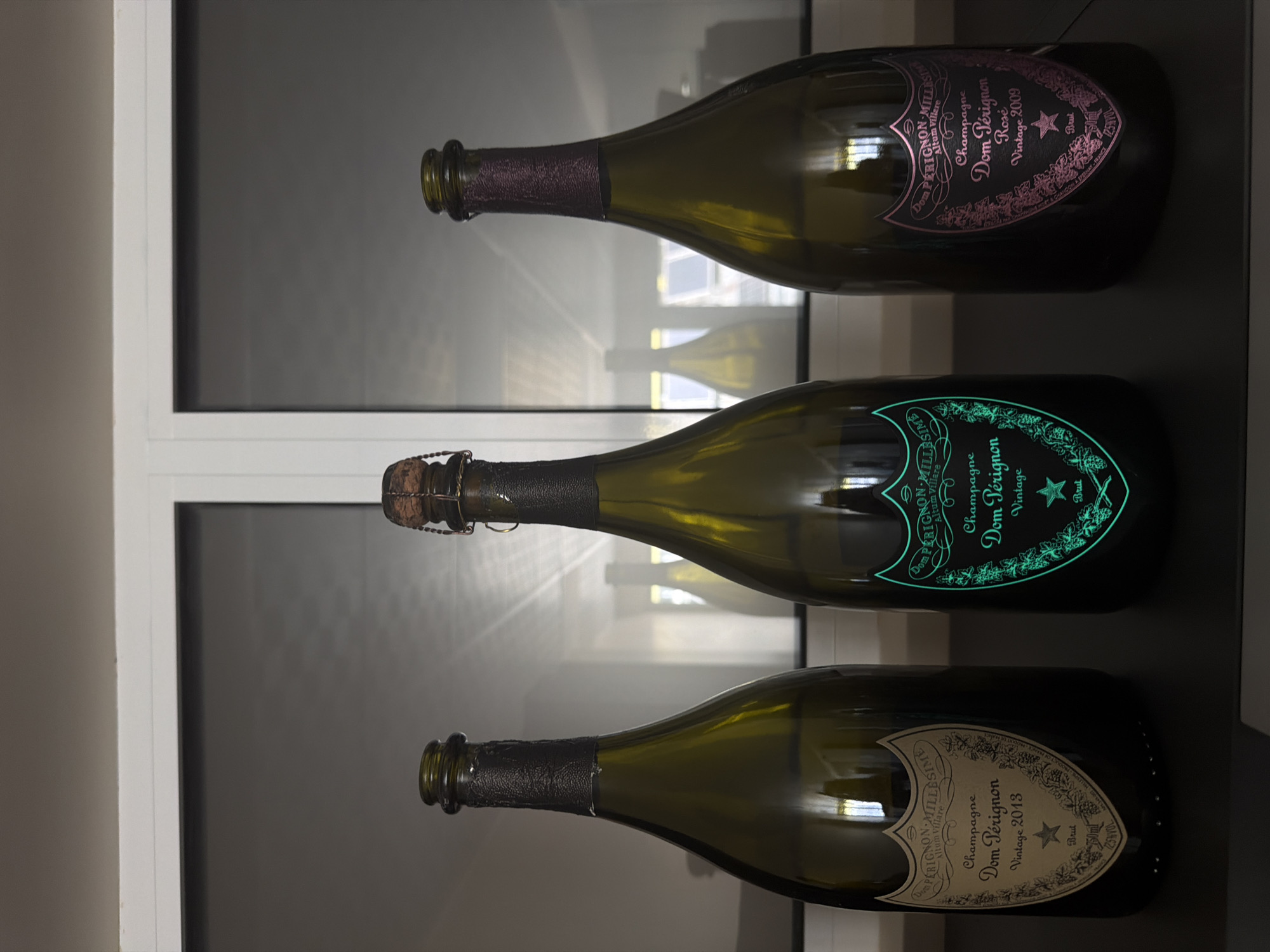 Three bottles of Dom Pérignon champagne are displayed on a dark surface in front of a window with white mullions and frosted glass panes. The bottles are made of dark green glass and have foil capsules. The leftmost bottle has a light-colored label with text and decorative elements. The middle bottle has a neon green label with glowing text and an outline of foliage and a star. The rightmost bottle has a dark pink or purple label with text and decorative elements. The lighting in the scene is dim, and there are reflections of the bottles visible on the surface in front of them. The overall setting suggests an indoor display, possibly in a home or a bar. The location of Dolno Ablanovo, Bulgaria is not discernible from the image.