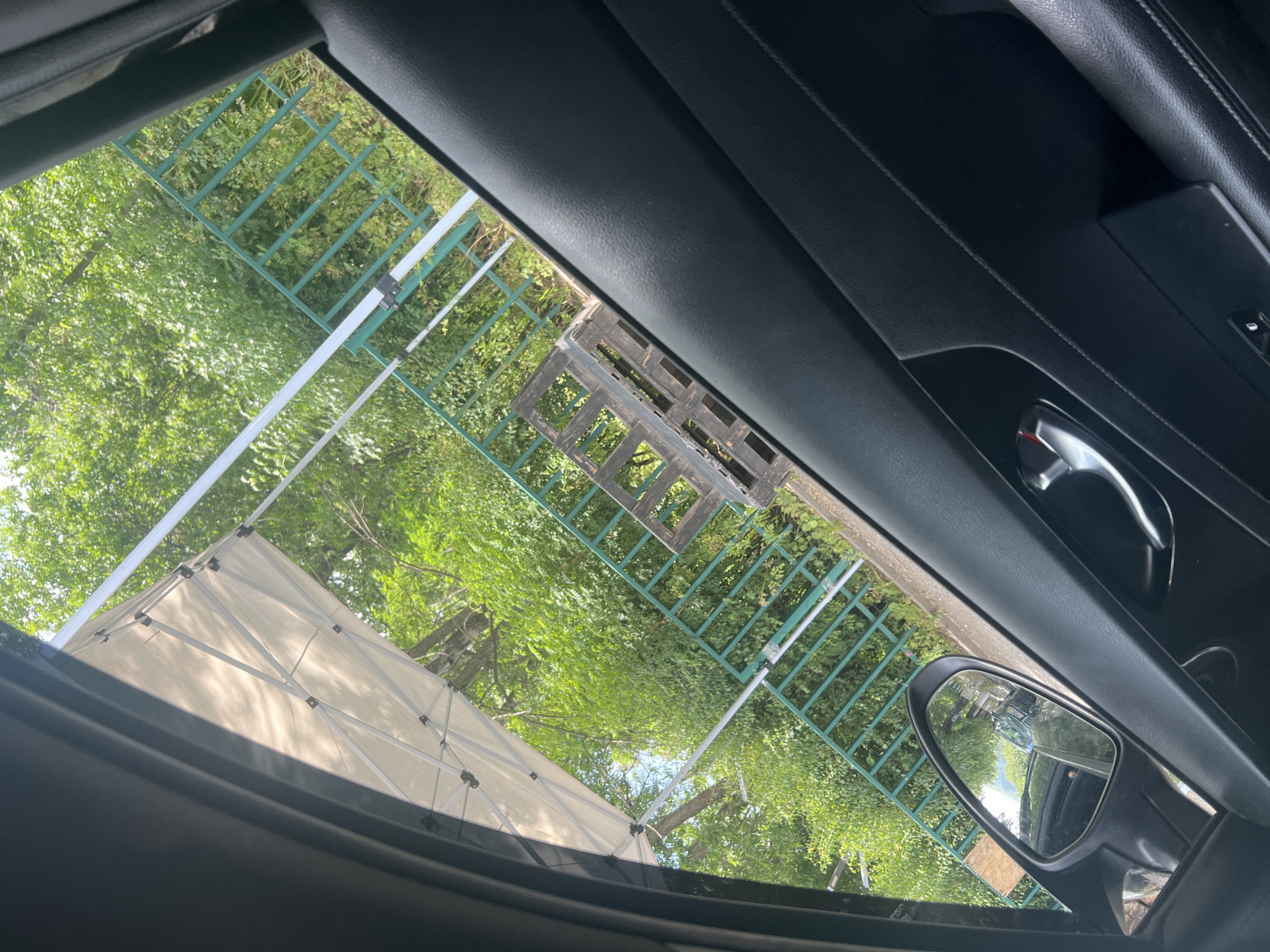 The image is taken from inside a car, looking out through the passenger-side window and partially through the sunroof. The perspective is angled, with the car's interior, including a black leather door panel and chrome handle, dominating the foreground on the right. To the left, a side mirror reflects a road and other vehicles, suggesting the car is in a parking area or on a street.
Outside the car, a white canopy tent is visible, supported by a metal frame. Beyond the tent, there is a dense green backdrop of trees and foliage. A green metal railing runs horizontally across the scene, separating the foreground from the background. Behind the railing, a wooden pallet is positioned on the ground. The scene appears to be outdoors, possibly in a park or a recreational area, given the presence of the canopy and greenery. The lighting suggests it is daytime, likely a sunny day given the bright highlights on the canopy and the overall vibrancy of the green foliage. The environment appears natural and somewhat unkempt, with some undergrowth visible near the railing and pallet.
While the image does not definitively place the scene in Vanadzor, Armenia, the query implies this location. Assuming this context, the scene depicts an outdoor setting within or near the city. The presence of the car and the urban-style railing suggests a developed area, possibly a public space or a roadside location. The wooden pallet could indicate a temporary setup, storage, or a remnant of past activity. The overall impression is one of an ordinary outdoor scene, likely captured during a stop or a drive through the city.