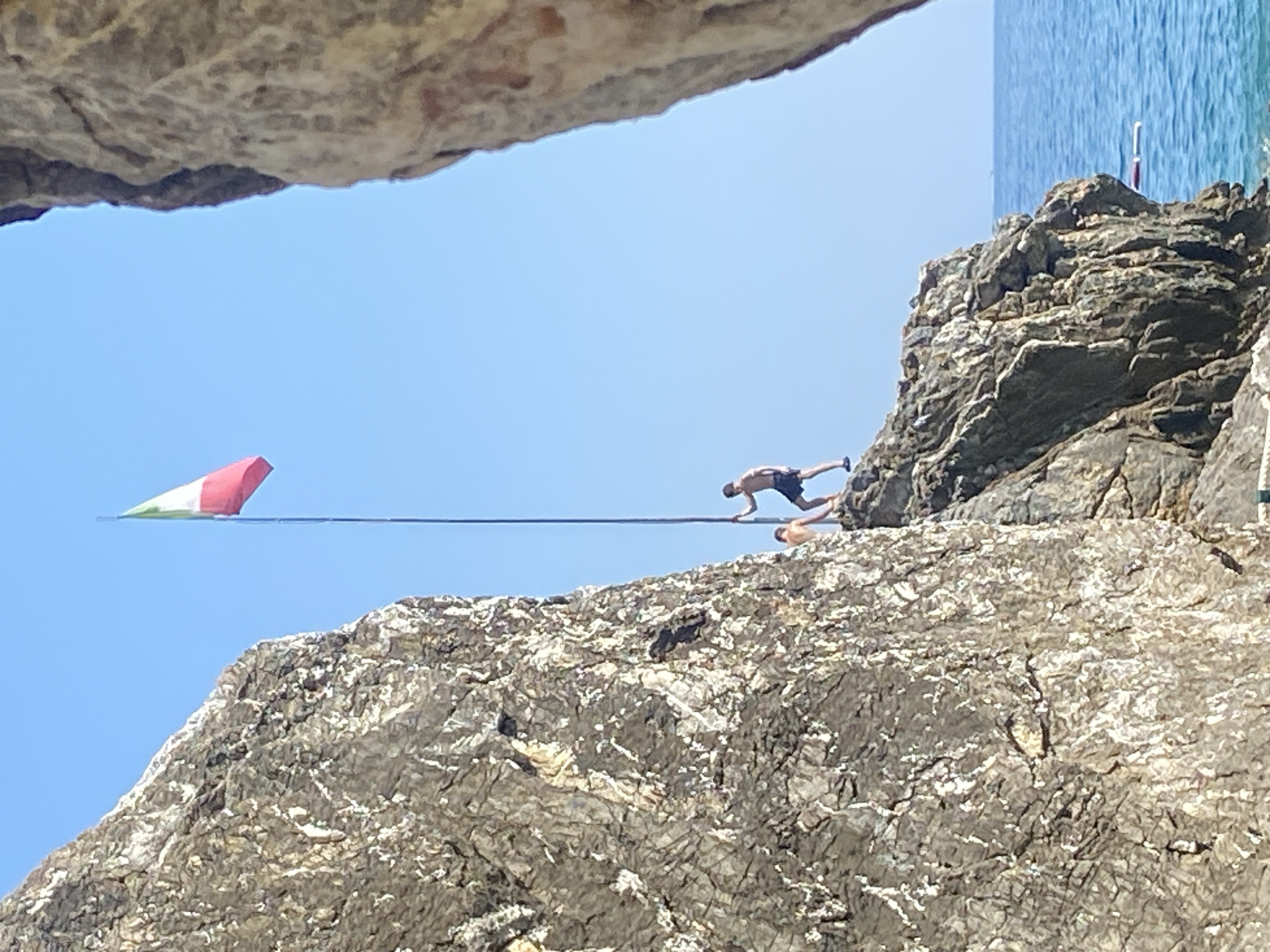 A group of two young men are standing on a rocky cliff overlooking the sea, one is holding a flagpole with an Italian flag. The rock face is rugged and textured, with clear blue sky above and the ocean below. One man is wearing black shorts and the other is shirtless. The scene appears to be a recreational area, possibly a beach or swimming spot. The location is the city of Prijevor, Montenegro.