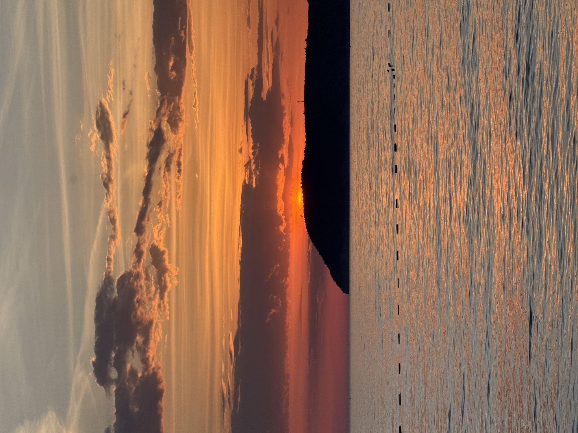 The image depicts a sunset scene over the Adriatic Sea, near Bar, Montenegro. The sky is filled with striated clouds, colored in hues of orange and pink by the setting sun. A dark, indistinct landmass, likely a mountain or coastline, is silhouetted against the sun. In the foreground, the water reflects the colors of the sky. A series of dark posts are evenly spaced in the water, creating a line that stretches into the distance. Several small birds can be seen resting on one of the poles.