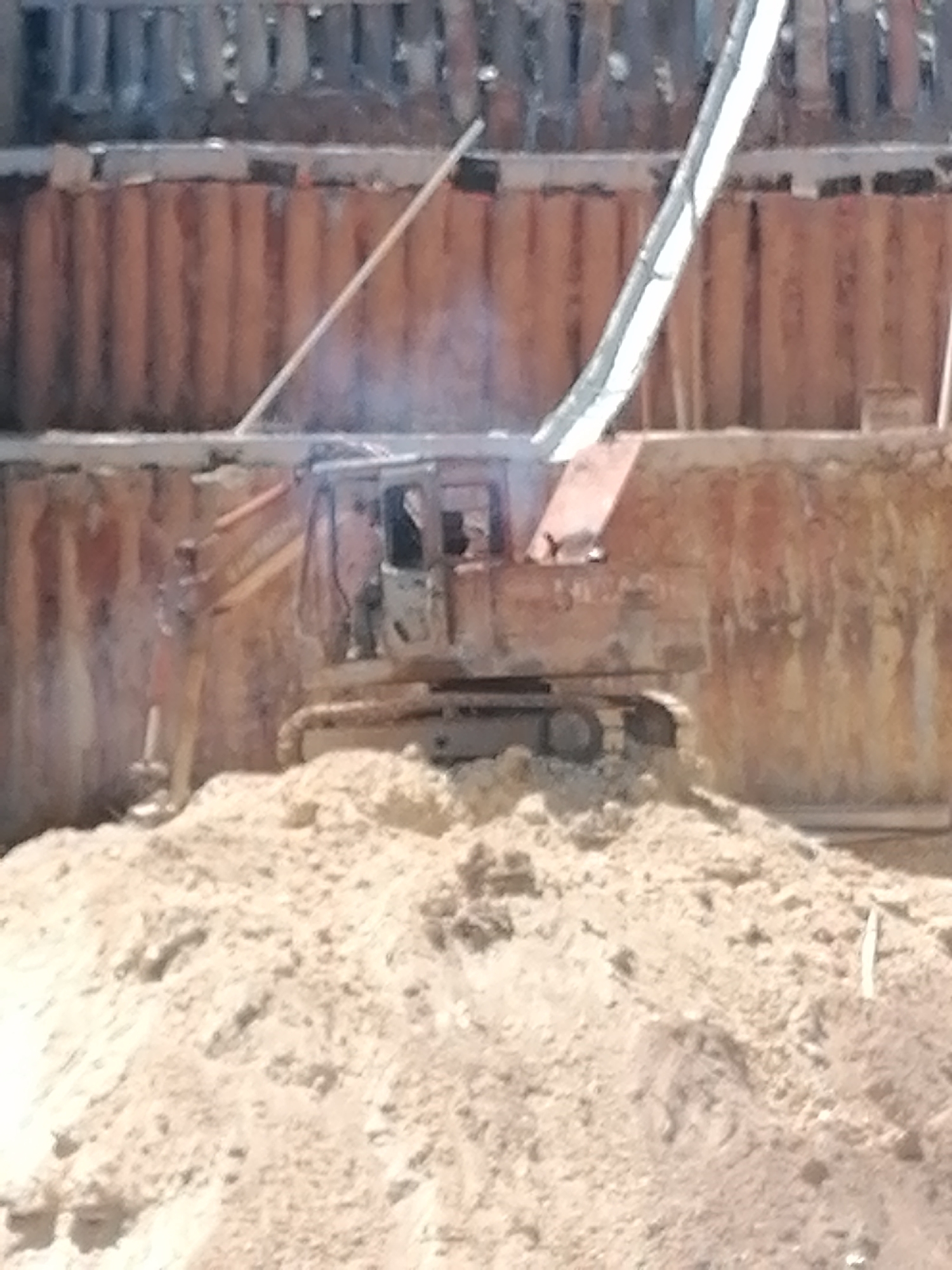 This image depicts a construction site in Da Lat, Vietnam. A large excavator, possibly an older model, is partially buried in a mound of soil. The excavator has an orange and white color scheme. The background shows a retaining wall made of corrugated metal sheets, topped with wooden planks. A metal pipe or conduit runs diagonally across the upper portion of the image, partially obscuring the retaining wall. The overall scene suggests ongoing excavation or earthmoving work.