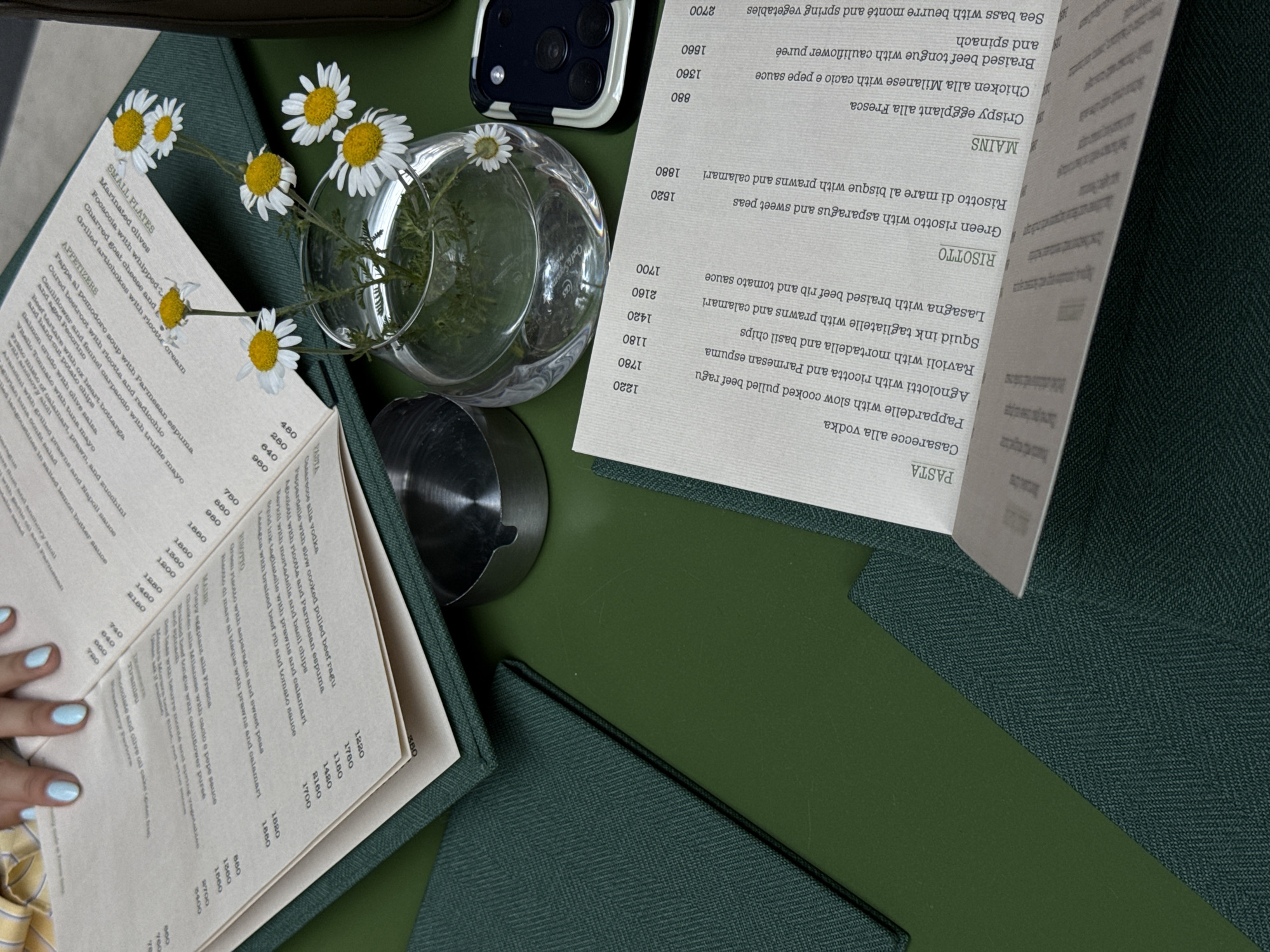The image shows a table setting in a restaurant. Two menus are visible, one on either side. There are daisy flowers in a transparent vase on the table. A phone is placed near the vase. There is a person with light blue nail polish touching one of the menus. The menu lists various dishes, including pasta, risotto, and mains, with corresponding prices. The image does not provide any information about the location or the city of Belgrade, Serbia.