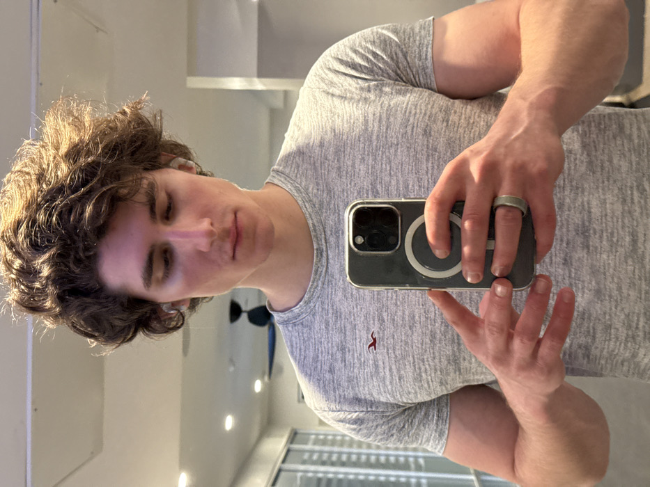 A young adult male, with brown, curly hair and fair skin, is depicted in a mirror selfie taken indoors. He wears a grey marl short-sleeved t-shirt featuring a small red Hollister seagull logo on the left chest. White wireless earbuds are present in both ears. He holds a silver-grey iPhone, enclosed in a clear, MagSafe-compatible case, with both hands at chest level to capture the image. A silver-grey ring is worn on the ring finger of his left hand. His gaze is directed downwards towards the phone. The background, a reflection in the mirror, reveals an interior space with a white ceiling containing multiple recessed light fixtures and a dark-colored ceiling fan visible in the upper left quadrant. A large glass partition or window with vertical elements is seen on the left, while a plain light-colored wall is on the right. The location is an indoor establishment within Los Angeles, California, United States of America.