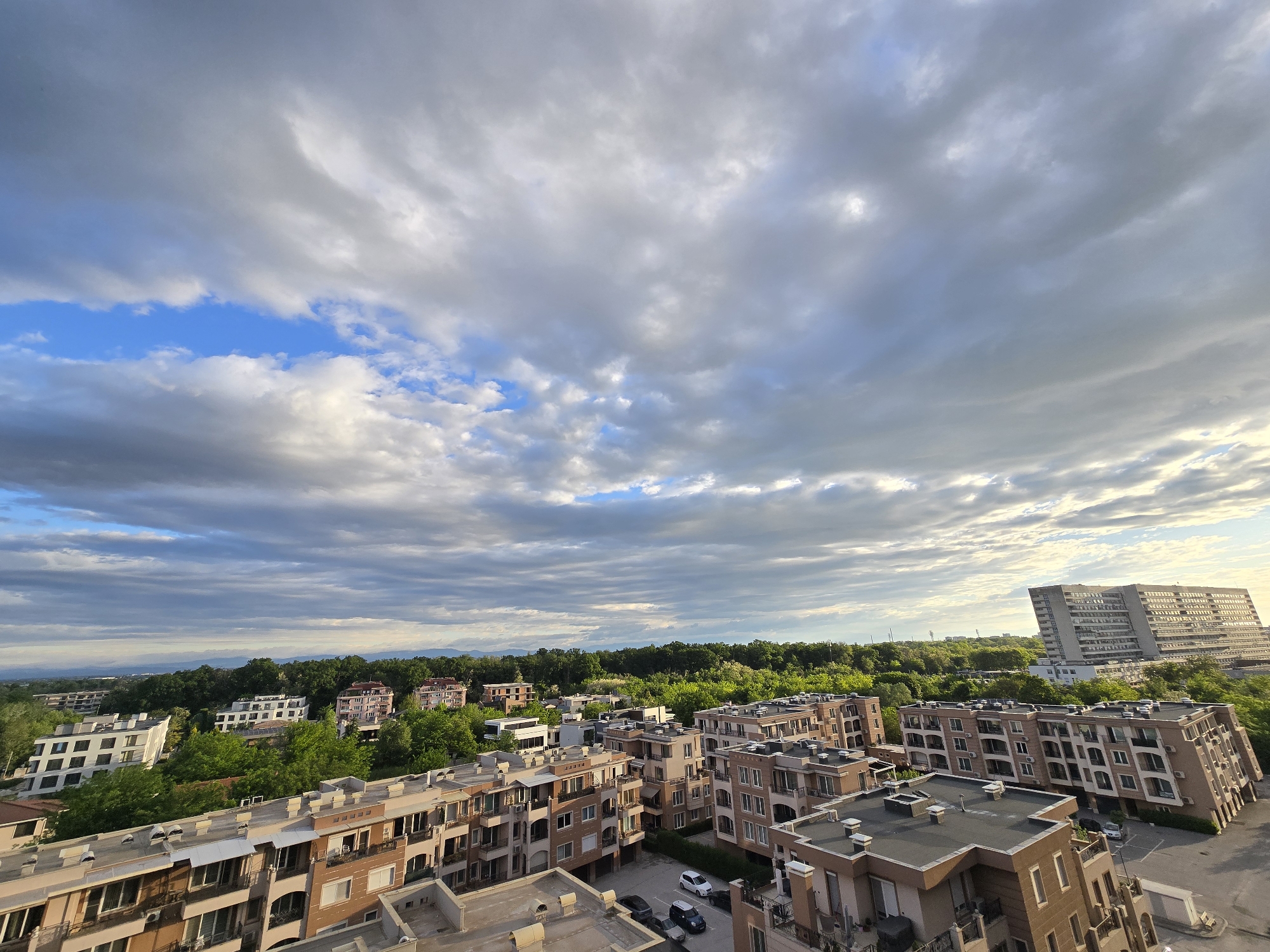 The image depicts a cityscape under a sky filled with predominantly grey clouds interspersed with patches of blue sky. The buildings are a mix of residential complexes with light brown and beige facades, some with visible balconies. In the background, there are trees that appear green and lush. A large, tall building stands out on the right side, casting a subtle, warm glow. There are several vehicles parked around the residential buildings. The overall impression is a view from a high vantage point, possibly a rooftop or upper-story window, with a wide view encompassing the city and its surrounding environment.