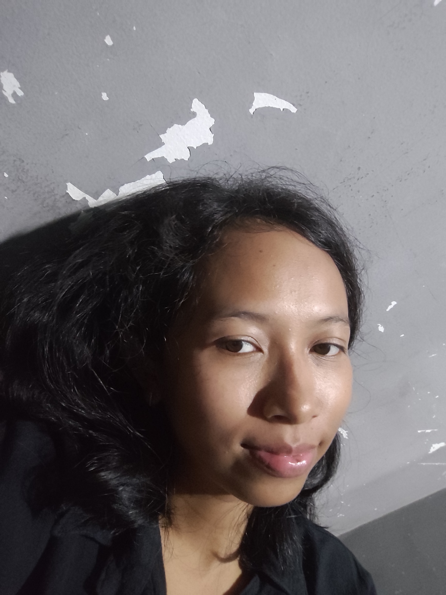 A close-up, medium shot shows a young woman's face, taken from a low angle. The woman has dark, wavy hair that is slightly messy. Her skin is smooth and slightly oily, with a warm undertone. She is wearing a black top. Her lips are full and glossy, with a slight smile. Her eyes are dark brown and slightly downcast. The background is a textured gray wall with peeling white paint. The lighting is dim, casting a shadow on the left side of her face. The scene appears to be indoors, possibly in a residential setting. The location is Unknown, Unknown.