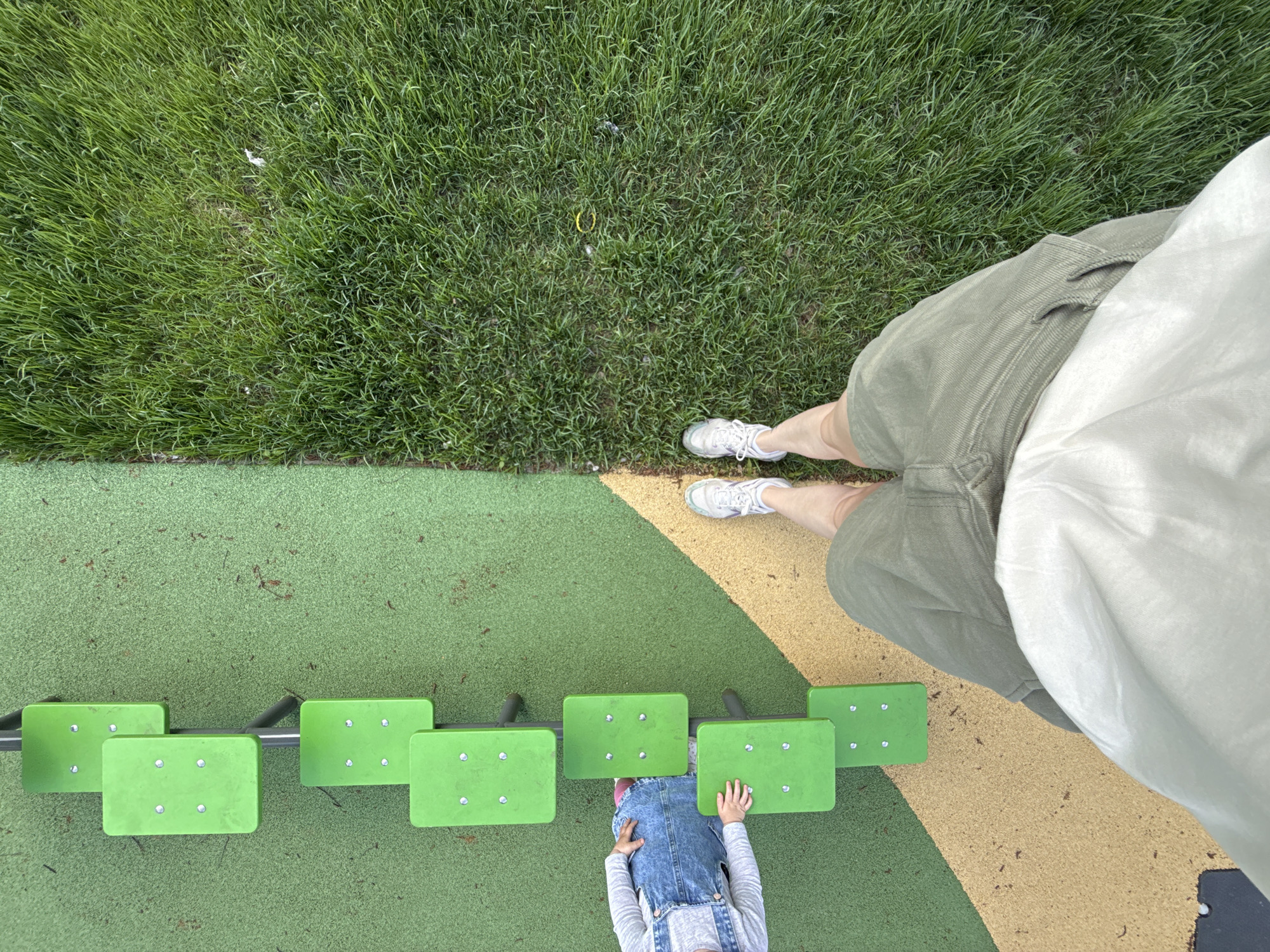 This image appears to be taken from a high, possibly first-person, perspective. The scene depicts a section of what appears to be a playground or recreational area. Part of the scene includes grass on the top side of the image. Below the grass is a section of playground floor composed of green, beige, and yellow sections. The floor holds play equipment that consists of a series of six elevated green stepping stones, possibly a balance beam of sorts. A child is shown reaching across the play equipment. The child wears a light grey shirt and jean overalls. Adjacent to the playground on the right side of the image, an adult stands in khaki shorts and a white shirt. The adult is wearing white shoes. The location could be Moscow, Russia.