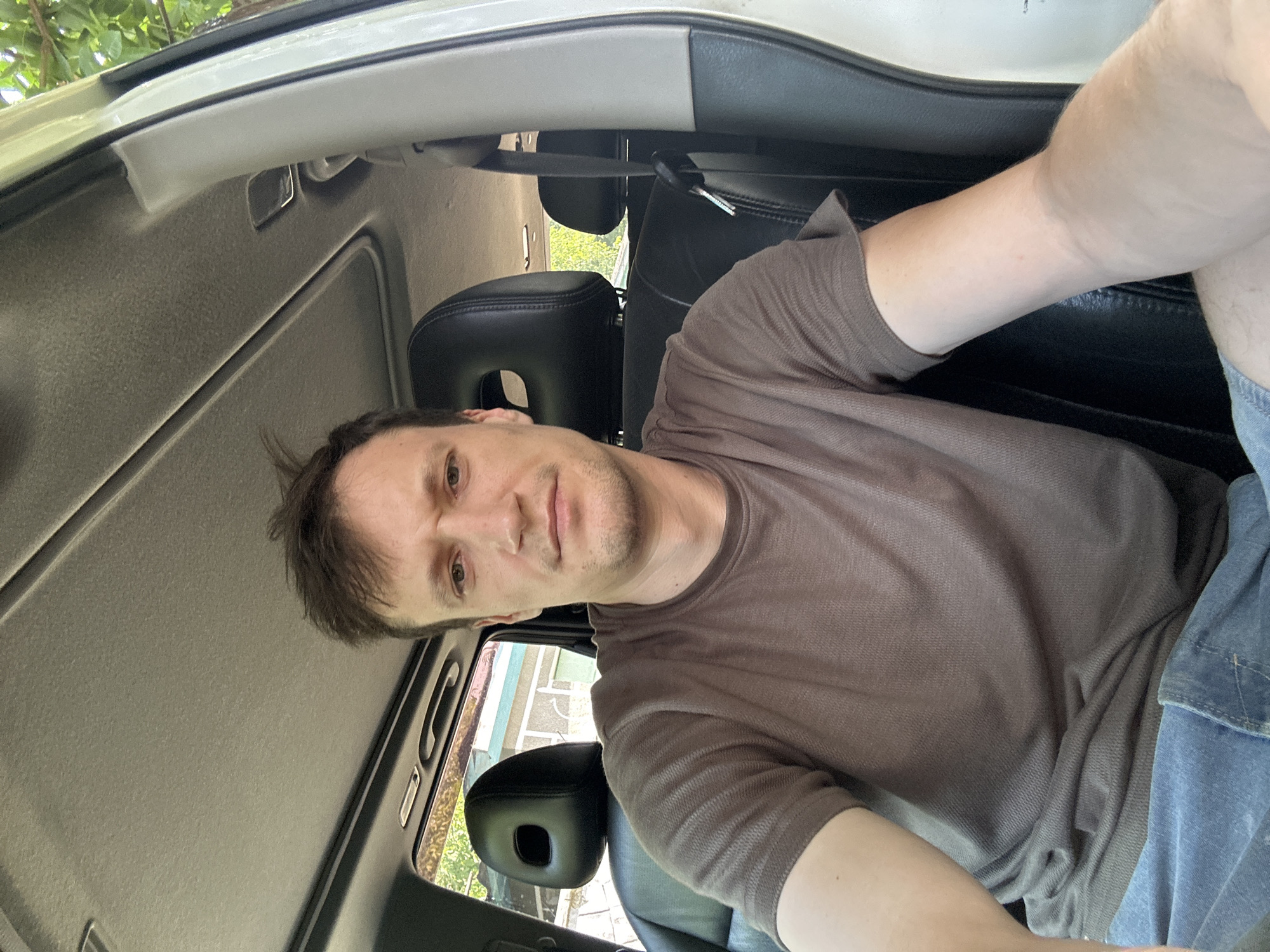 A man is sitting in the driver's seat of a car. He is wearing a brown t-shirt and blue jeans. The car is parked outside, and trees and a building can be seen in the background. The man appears to be looking at the camera. The image was taken in Makiv, Ukraine.