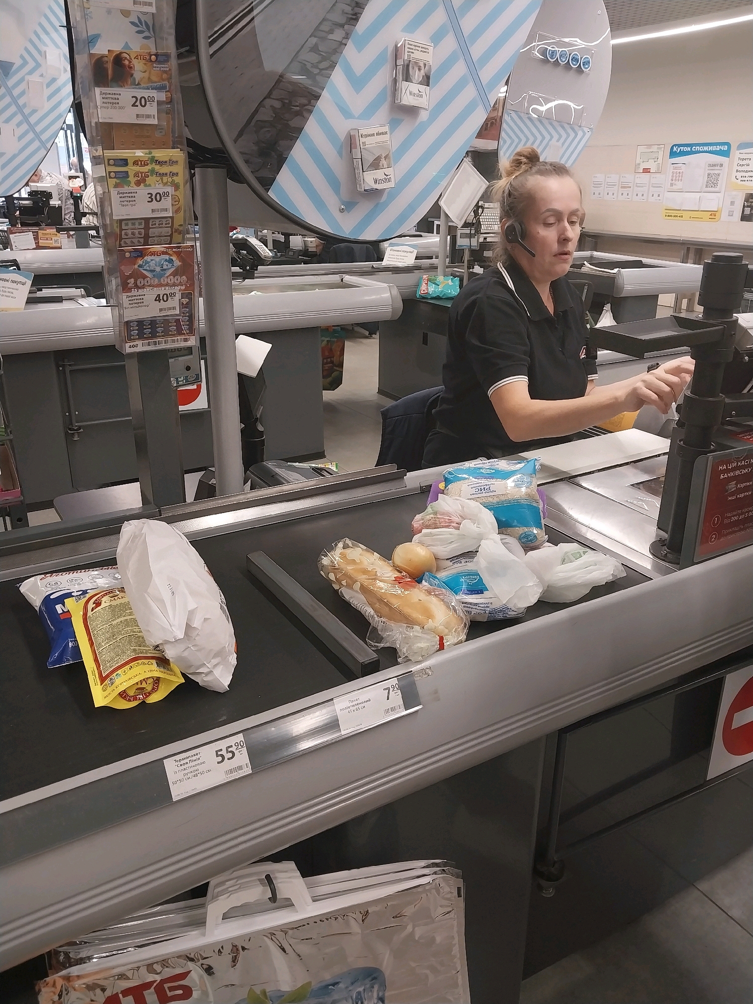The image shows a cashier at a supermarket in Dnipro, Ukraine, processing items on a conveyor belt. The cashier, a woman with a headset, is wearing a black polo shirt with a red logo. Several grocery items, including bread, onions, and packaged goods, are on the belt. In the background, there are displays of lottery tickets and cigarette packs. The supermarket environment appears typical, with a focus on the checkout area.