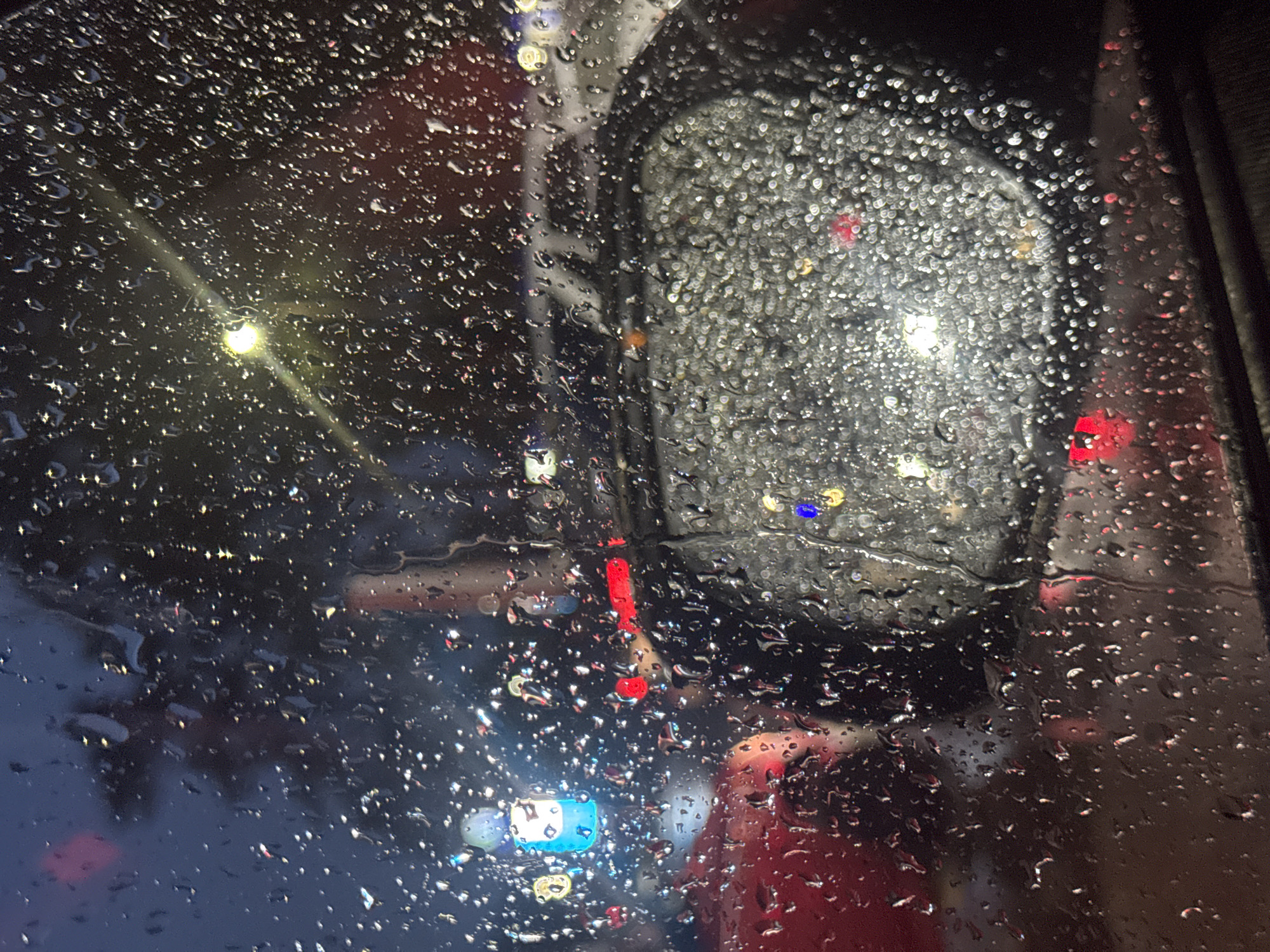 The image depicts a view through a rain-streaked window, likely from inside a vehicle, at dusk or night in the city of Chaikovsky, Russia. The foreground is dominated by numerous water droplets of varying sizes adhering to the glass, distorting the scene beyond.
In the middle ground, a car's side-view mirror is visible, also covered in water droplets which create a bokeh effect of blurred lights. Reflected in the mirror are various colored lights, including bright white, yellow, and red, suggesting streetlights, traffic lights, or illuminated signs.
Beyond the mirror and the window, the background shows a street scene with out-of-focus vehicles and buildings. Red lights, possibly from taillights of cars or traffic signals, are prominent. A bright blue light is also visible, possibly from a sign or building illumination. The overall atmosphere is dark and moody, characteristic of a rainy evening in an urban environment. The presence of reflections and the nature of the lights suggest a typical city setting.