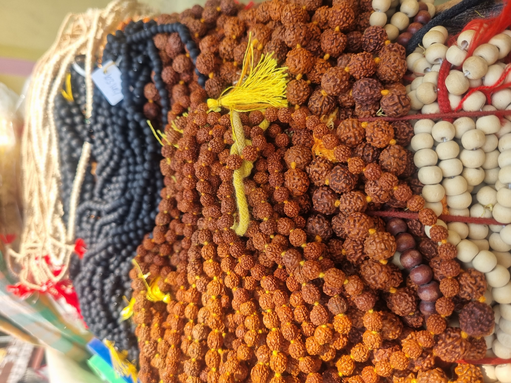 The image displays a close-up, elevated view of various religious prayer beads (malas) arranged in piles, likely at a market stall or shop in Puttaparthi, India.
The dominant subjects are numerous strands of reddish-brown Rudraksha beads, characterized by their textured, multi-faceted appearance. These are strung on yellow thread, with several strands featuring bright yellow tassels. They occupy the majority of the foreground and midground.
Behind and partially beneath the Rudraksha beads, a large quantity of smooth, uniformly black, spherical beads is visible. A white rectangular price tag, partially legible as "INR 50/-", is attached to one of these black bead strands.
In the bottom portion of the frame, two distinct sections of light-colored, smooth, round wooden beads are visible, separated by thin red and black cords.
The background is out of focus, showing blurry details of lighter-colored woven material (possibly garlands) and indistinct patches of green and blue fabric. No individuals or specific actions are depicted within the scene. The overall environment suggests a retail display of devotional items.