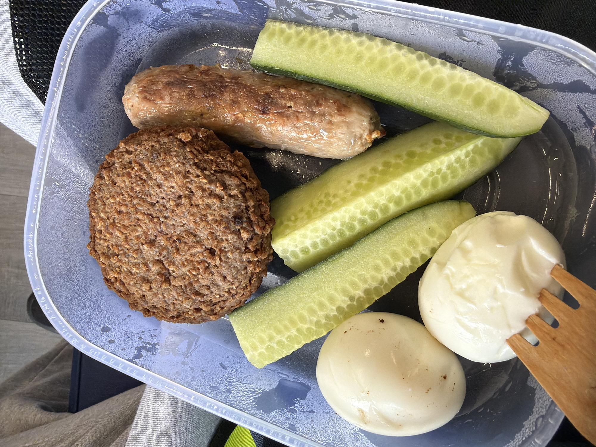 A top-down shot shows a partially filled, rectangular, transparent plastic container with a black interior base. The container holds a selection of food items: a browned, circular meat patty; a cooked sausage with a light brown casing; four sticks of cucumber, uniformly cut; and two peeled hard-boiled eggs, one with a dollop of white creamy sauce. A wooden fork is partially visible, resting against one of the eggs coated in sauce. The background reveals details of what appears to be clothing or fabric, with the edge of a bag visible at the top left and green/yellow stripe. No information to locate the image within Connah's Quay, UK is contained within the image.