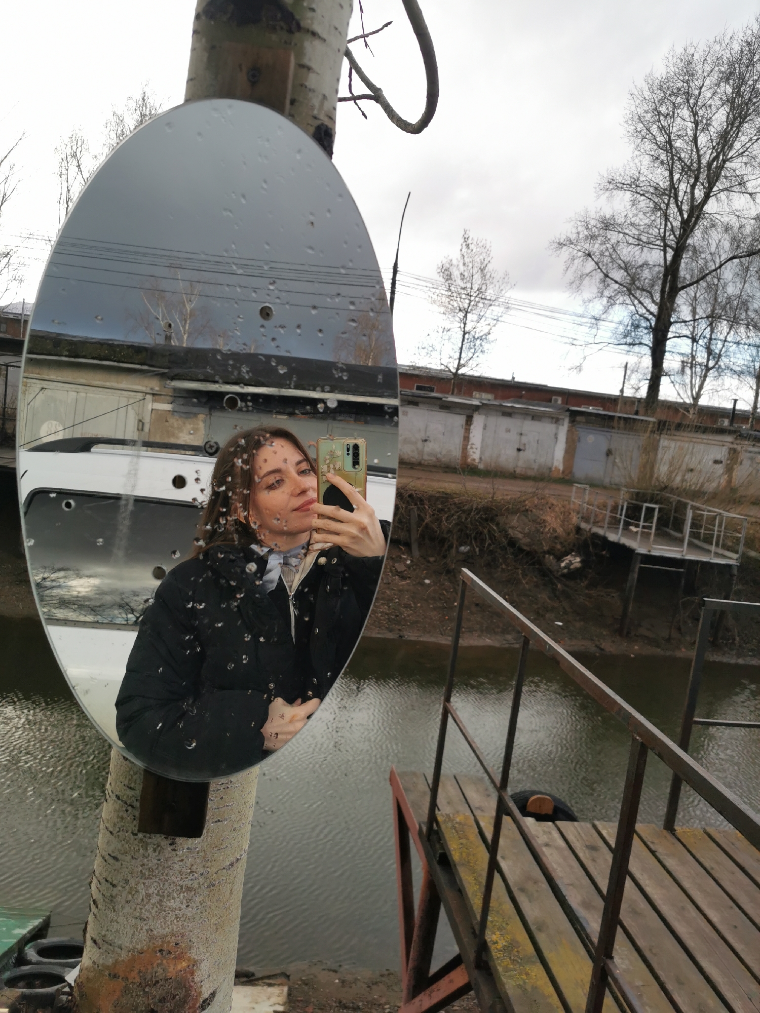 The image shows a woman taking a selfie in a mirror attached to a tree trunk. The mirror is oval and has raindrops on its surface. The woman is wearing a black puffer jacket, a light-colored shirt, and a scarf. She is holding a mobile phone in her hand. The reflection in the mirror shows part of a white vehicle.
Behind the mirror, the environment features a body of water, likely a river or canal, with a simple wooden pier or platform extending over it. On the opposite bank are a series of garages with a metal railing along the bank. The sky is overcast. The scene appears to be in an urban area.