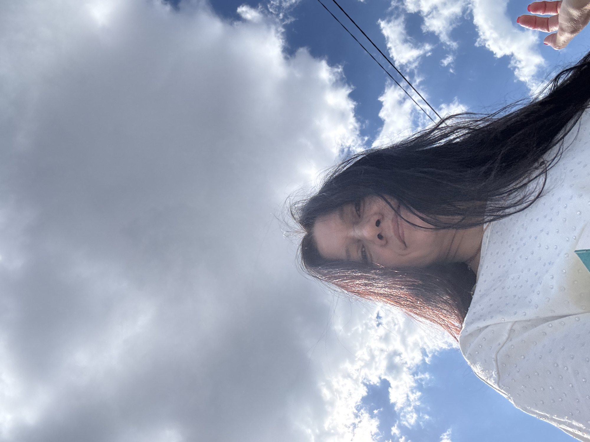A low-angle shot of a woman with long dark hair. She is wearing a white top. Her face is partially visible. In the upper right corner is a hand. The background consists of a partly cloudy sky and power lines. The location is Chaikovsky, Russia.