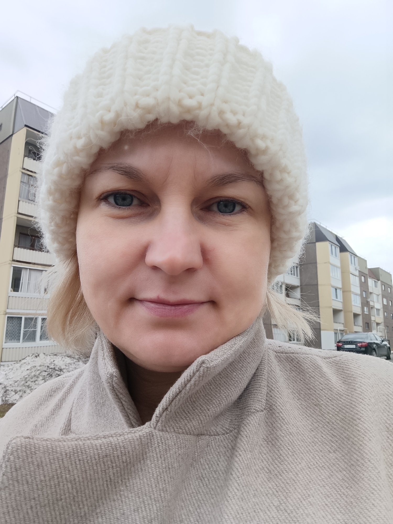A close-up, eye-level selfie depicts a middle-aged woman with fair skin and blue eyes. She is wearing a chunky, off-white knitted hat and a beige jacket with a high collar. Her blonde hair is visible underneath the hat.
The background reveals an urban setting with multi-story apartment buildings in neutral colors. A black car is parked in the distance. There is snow visible on the ground in a small patch. The sky is overcast.
Based on the buildings in the background, the location appears to be in Марковский, Russia.