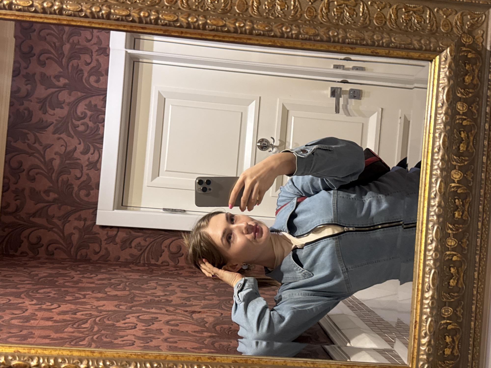 A young woman with light brown hair pulled back is reflected in an ornate gold-trimmed mirror, taking a selfie. She is wearing a light blue denim jacket over a cream-colored turtleneck. Her left hand is raised to her head, touching her hair. She is looking at the phone in her right hand. Behind her, a cream-colored door with raised panels is visible. The wall to her left is covered in patterned maroon wallpaper. The scene is indoors, likely in a bathroom or entryway, and the location is not identifiable as Shkotovo, Russia.