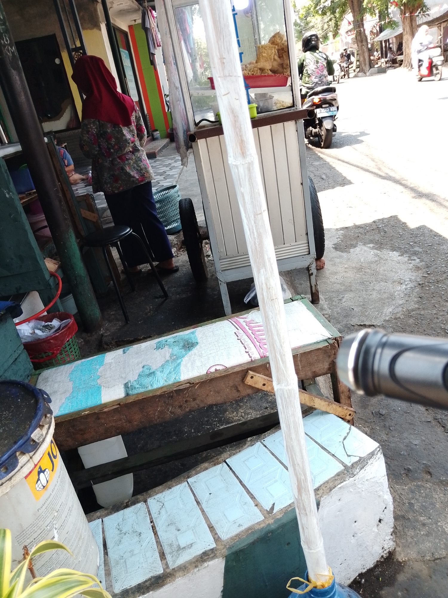 In Pasuruan, Indonesia, a woman wearing a red hijab and a floral-patterned shirt stands facing away from the camera. She is looking at a street vendor's cart which is selling fried food. Behind the cart, a man on a motorcycle, wearing a black helmet, rides past. The street is paved and lined with buildings and trees. The scene appears to be in a busy urban area.