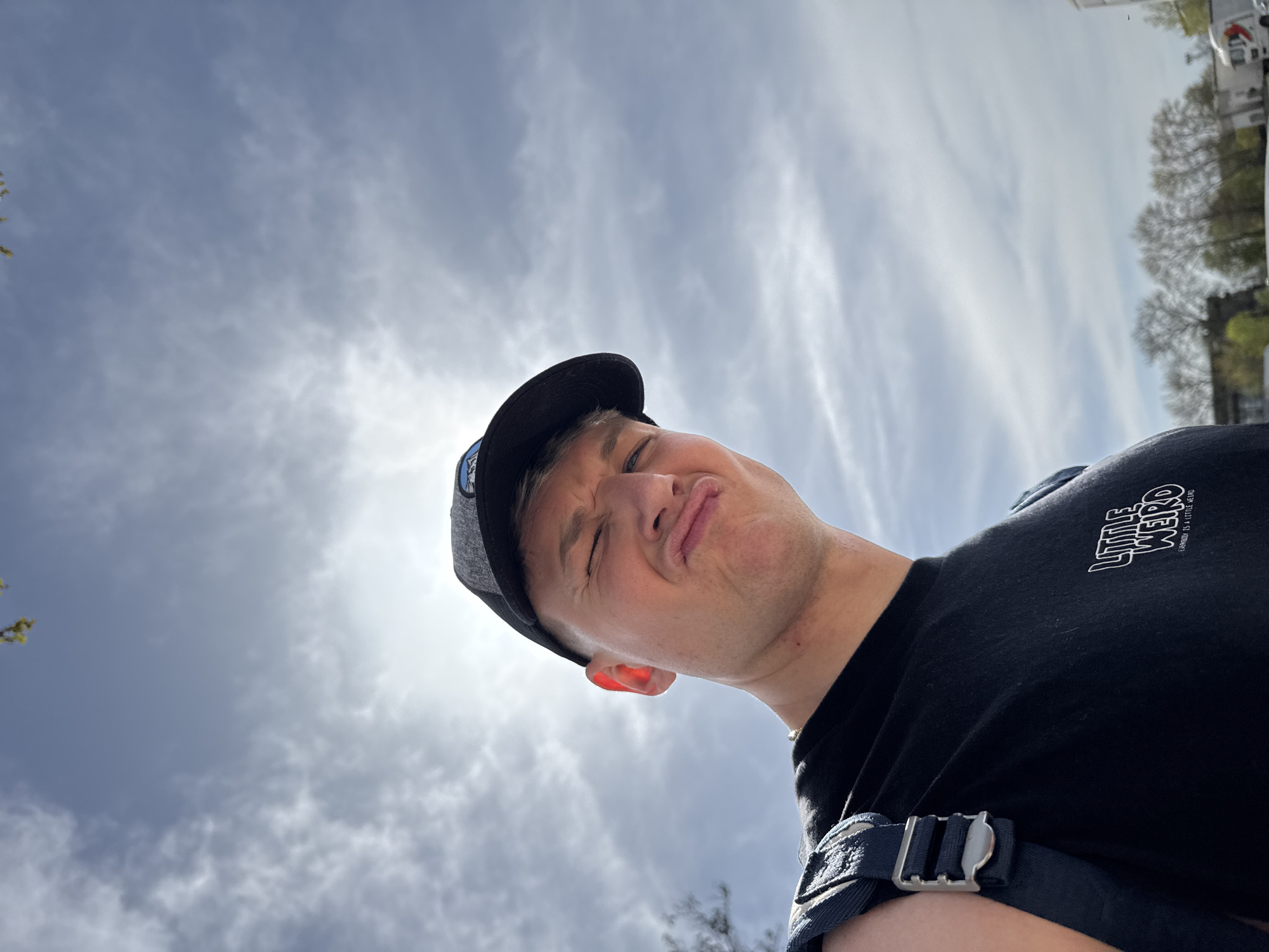 A low-angle outdoor portrait features a young man captured from below, looking down towards the camera with a playful, squinted facial expression and puckered lips. He is wearing a dark baseball cap and a black t-shirt. Bright sunlight, emanating from behind him, casts a strong glare in the upper sky and illuminates the edge of his ear, indicating daytime. The sky is partly cloudy, showing a mix of bright and darker cloud formations.
On the far left, light green foliage of a tree is partially visible, while on the far right, blurry distant structures, possibly buildings or vehicles, suggest an urban or suburban environment in Plovdiv, Bulgaria. A dark blue or black strap with a prominent silver buckle, likely part of a backpack or bag, is visible over his shoulder. The text "LITTLE WEIRD" and "EVERYONE IS A LITTLE WEIRD" is clearly printed in white on his black t-shirt. The overall scene conveys a casual and lighthearted moment.