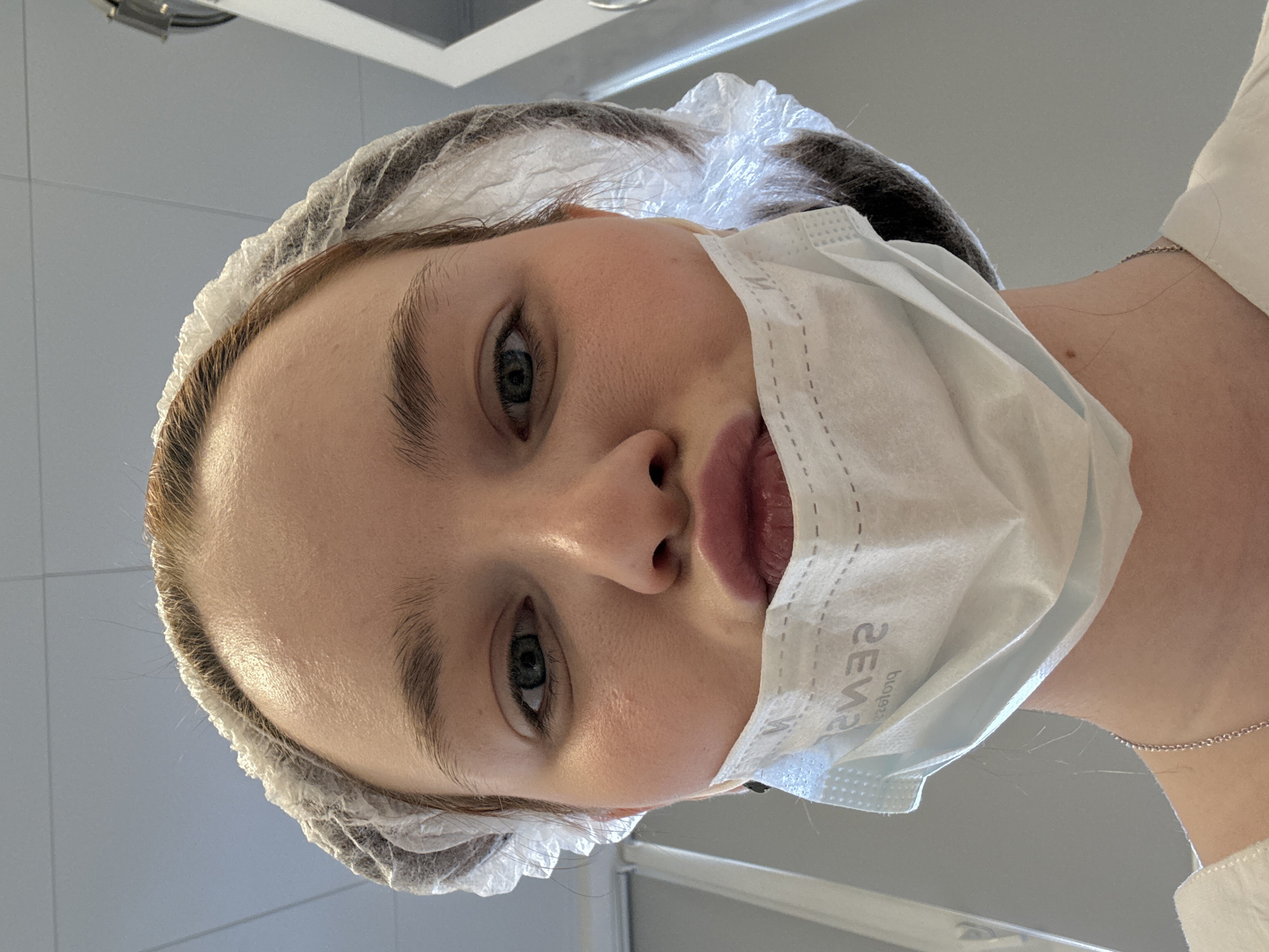 A person wearing a disposable hairnet and a face mask is seen from a low angle. The person has fair skin, light-colored eyes, and reddish-pink lips visible above the mask. The mask, which covers the lower part of their face, appears to be a standard surgical-type mask with pleats. The hairnet is white and covers the person's hair completely. The background appears to be a room with light-colored walls and potentially a medical or clinical setting due to the attire. The location is not discernible as Saint Petersburg, Russia from the available image.