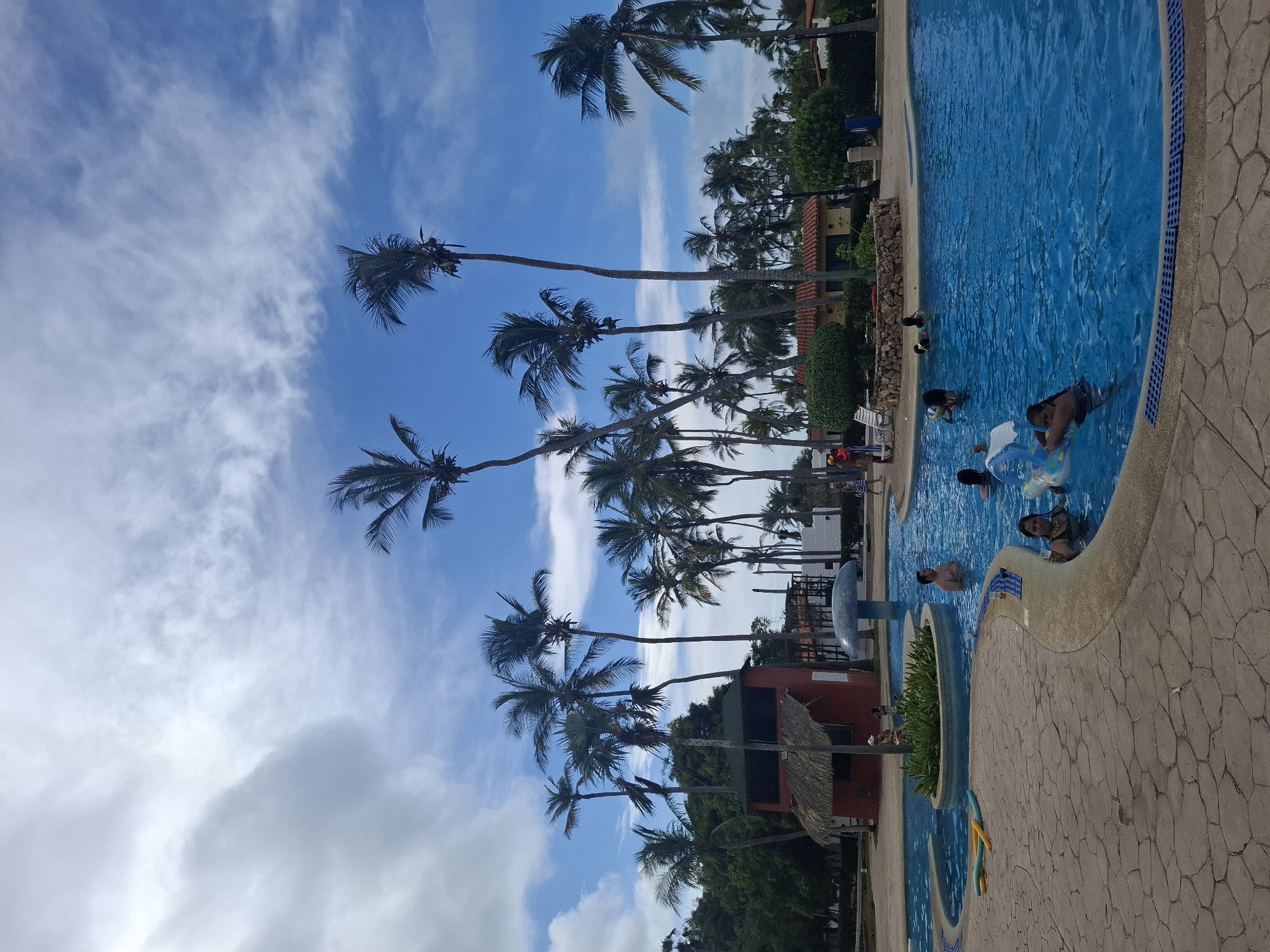 The image shows a swimming pool at a resort in Playa El Agua, Venezuela. Several people are in the pool, some with inflatable toys. Palm trees line the edge of the pool, with a blue sky and scattered clouds above. The pool has a textured stone surround. Buildings with orange tile roofs are visible beyond the palm trees.