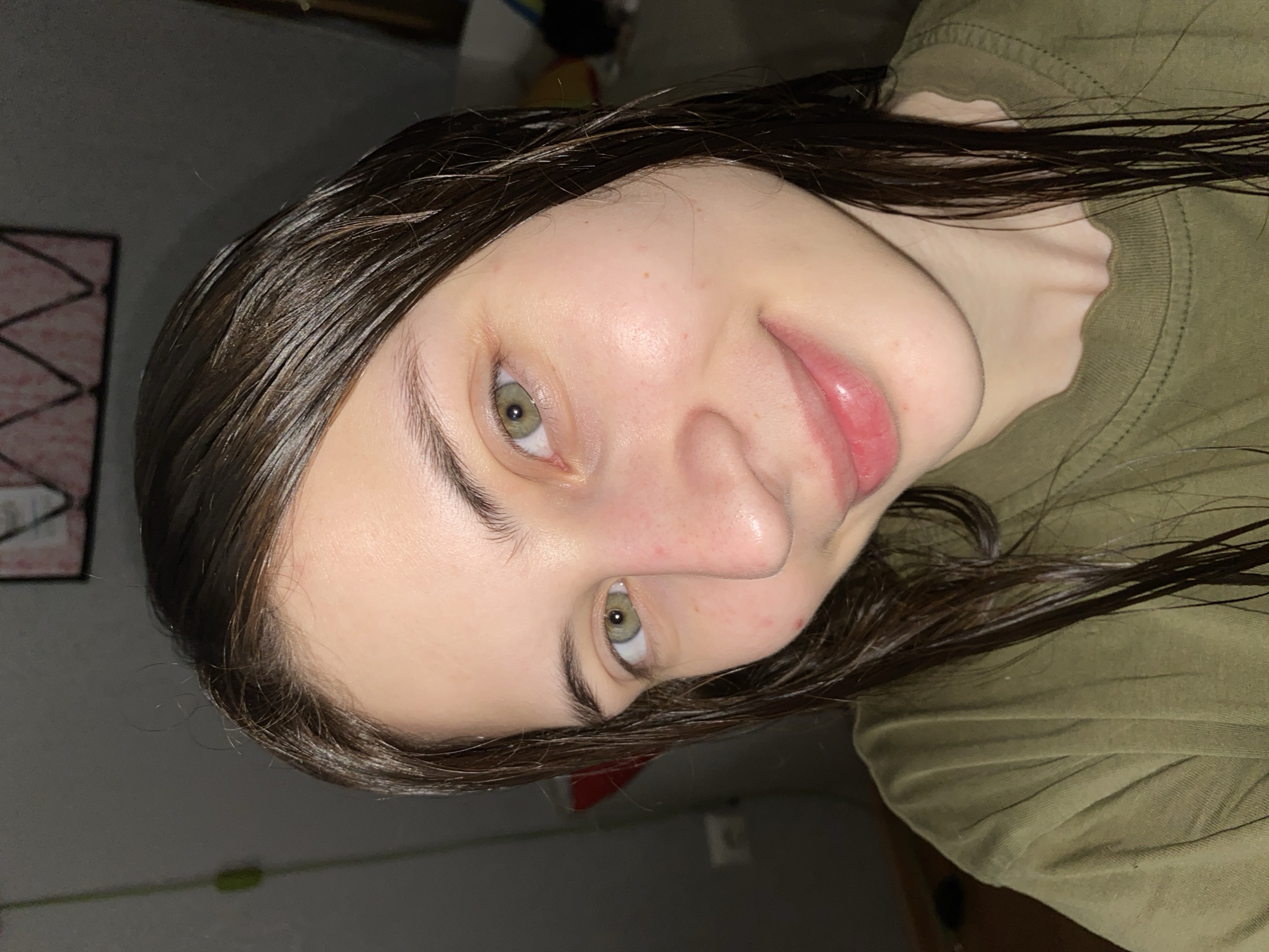 A close-up, slightly rotated, headshot of a young Caucasian woman with wet, dark brown hair. She has fair skin, green eyes, and a small amount of acne. She is wearing a drab green t-shirt. The background is a dimly lit interior with a blurry painting on the wall. There is no indication of this photo being taken in Votkinsk, Russia.