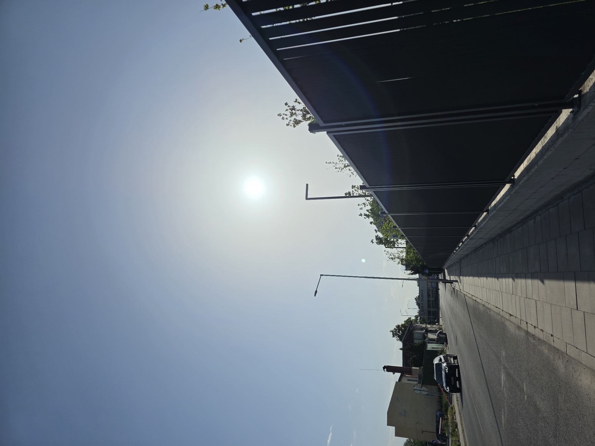 The image depicts a street scene in Plovdiv, Bulgaria. The sky is bright with the sun high in the frame. A long, dark structure with horizontal slats runs along the upper right of the image, possibly a covered walkway or building element. A paved road stretches into the distance, with a sidewalk on the right. A few cars are visible on the road. Streetlights line the street. Buildings of varying heights are seen on the left. Greenery is interspersed among the structures.